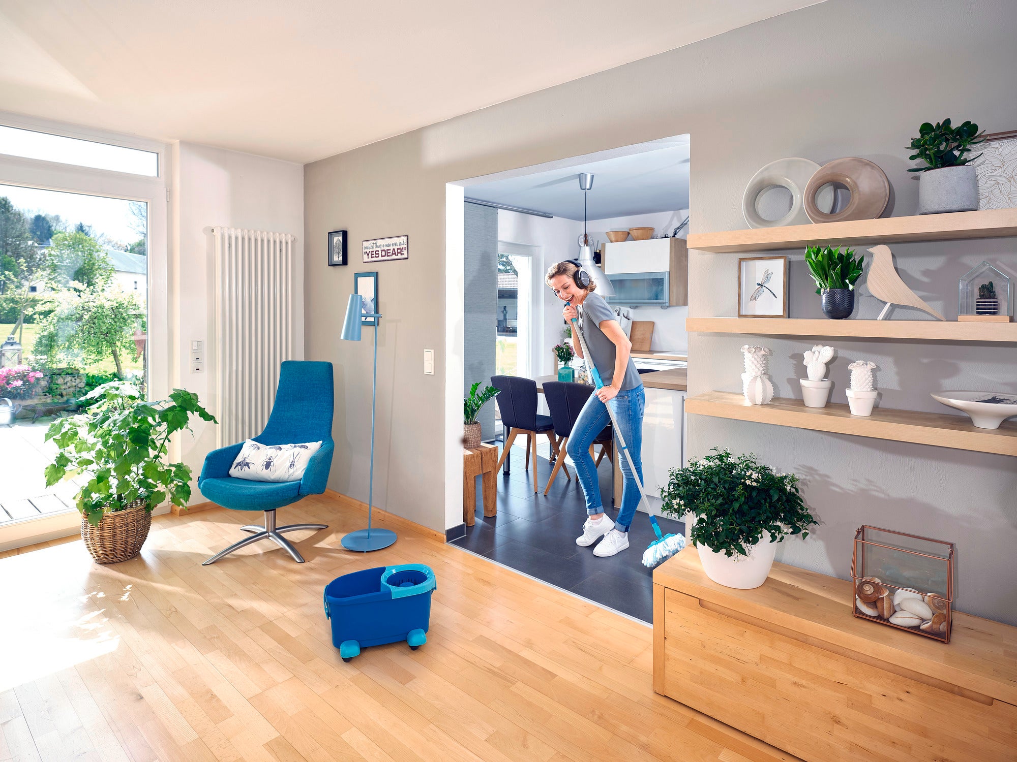A woman cleans the floor of a modern living room with the CLEAN TWIST Disc Mop Ergo. The room is bright and stylishly furnished with plants and decorations.