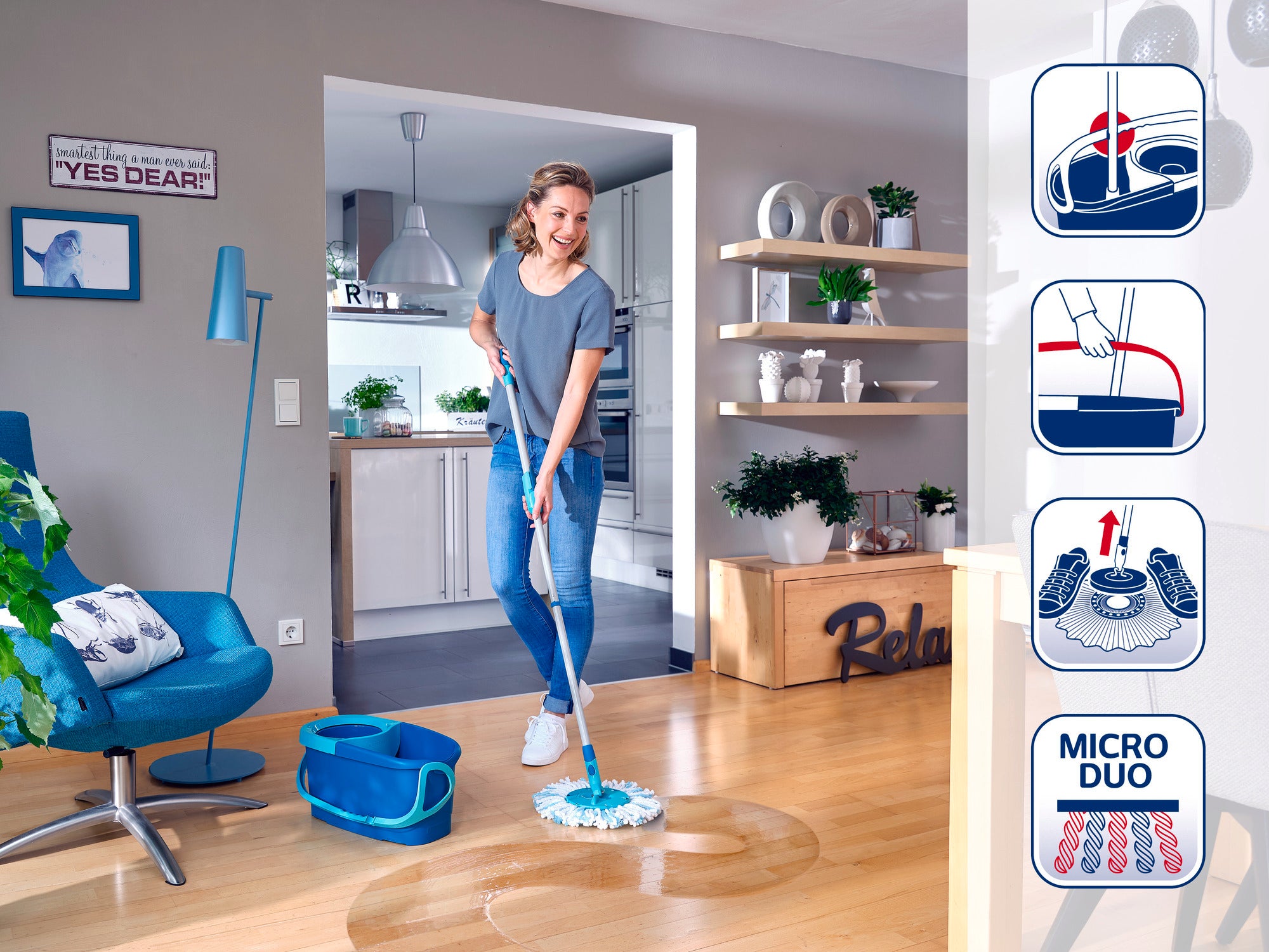 Kobieta myje drewnianą podłogę w nowoczesnym salonie mopem Leifheit. W tle widać otwartą kuchnię. Mop ma niebieskie wiadro i specjalną nakładkę z mikrofibry.