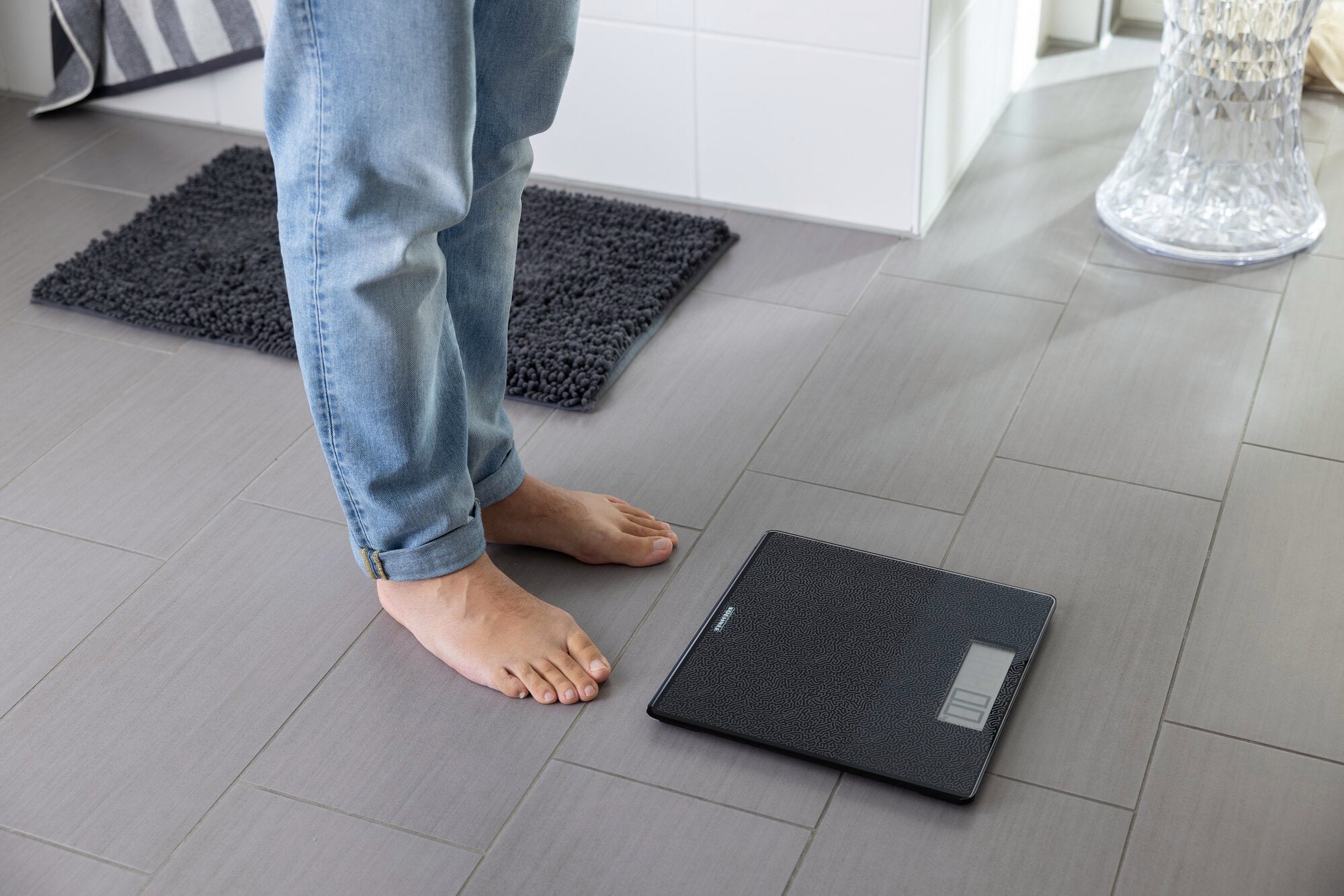 Een persoon staat blootsvoets op een badkamervloer naast een digitale weegschaal. De weegschaal heeft een plat en modern design, passend bij de stijlvolle omgeving.