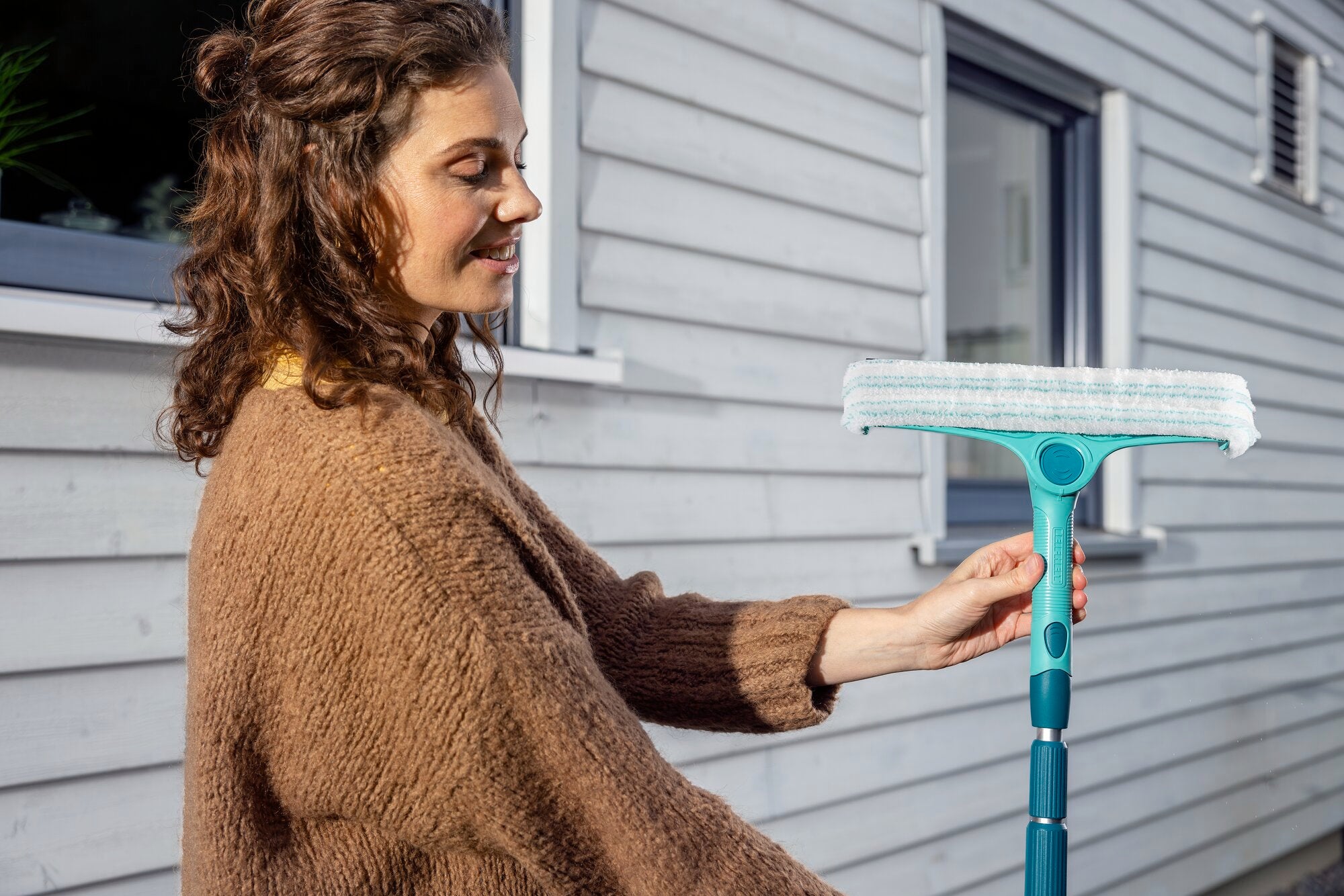 Kobieta używa teleskopowego uchwytu, który można regulować w zakresie od 145 do 400 cm, aby umyć okno. Trzonek ma turkusowy kolor i białą głowicę czyszczącą.