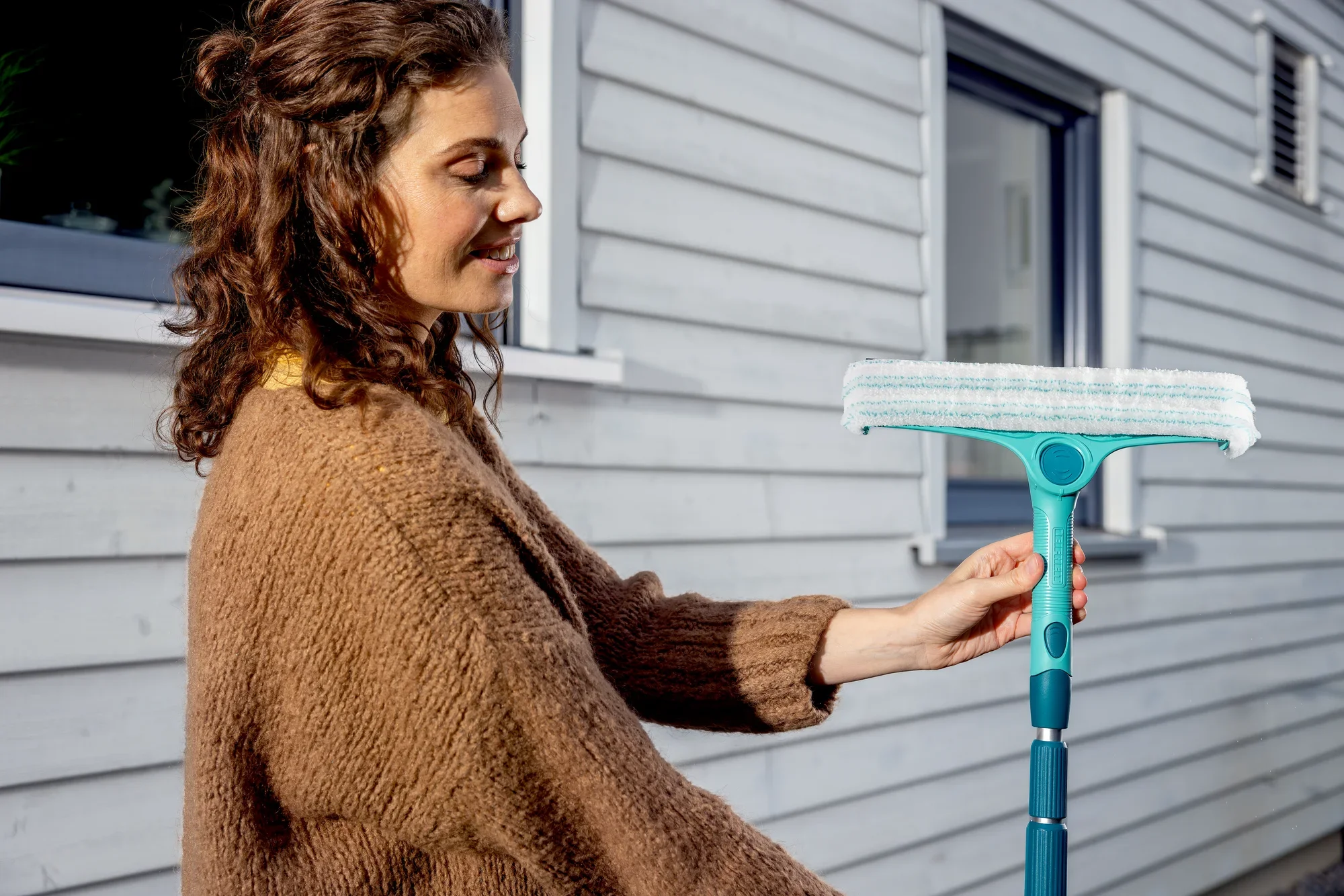 Kobieta używa teleskopowego uchwytu, który można regulować w zakresie od 145 do 400 cm, aby umyć okno. Trzonek ma turkusowy kolor i białą głowicę czyszczącą.