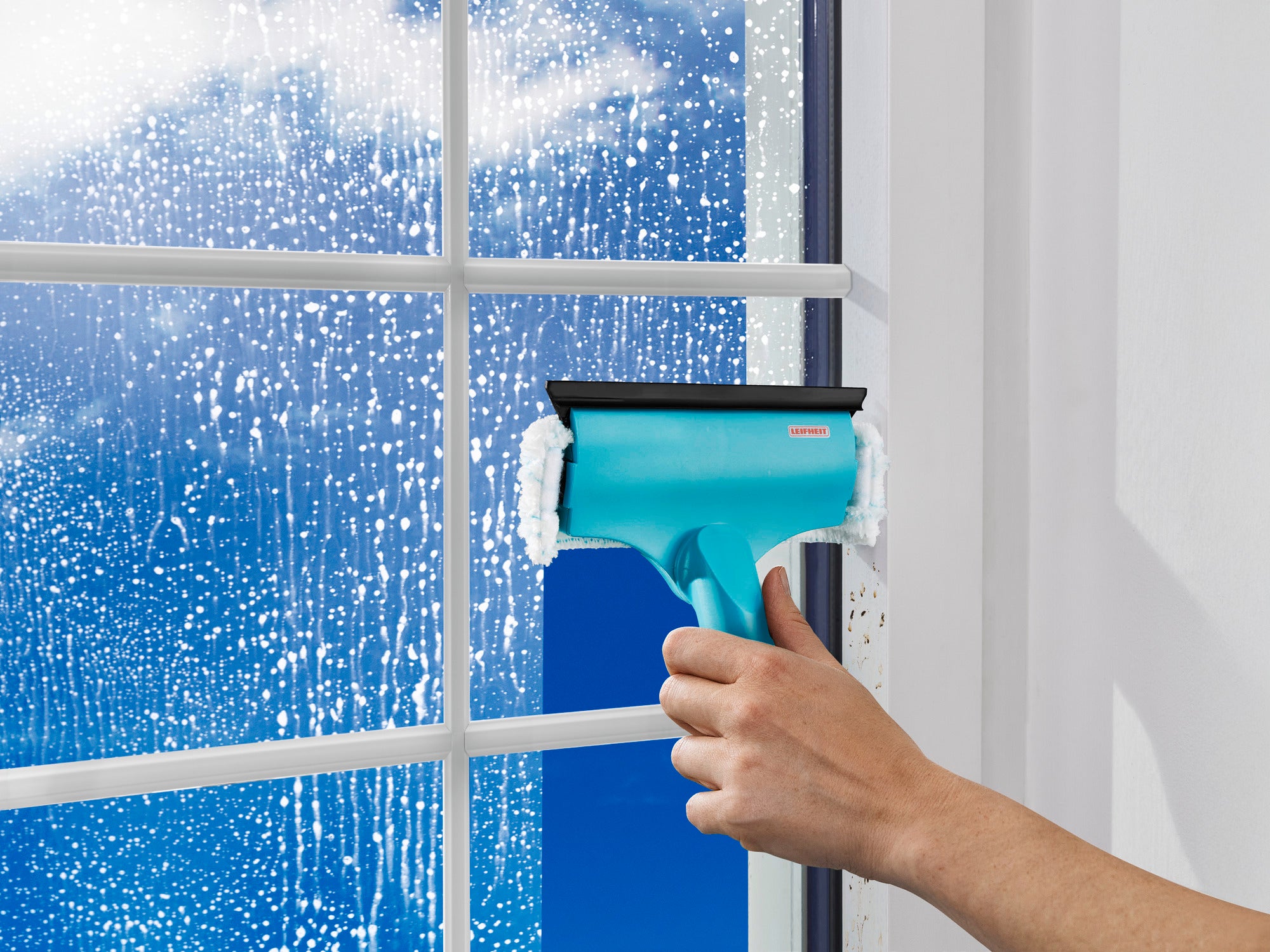 A 3-in-1 mini window and frame wiper is used to clean a window. The hand holds the turquoise wiper while water and foam are visible on the glass.