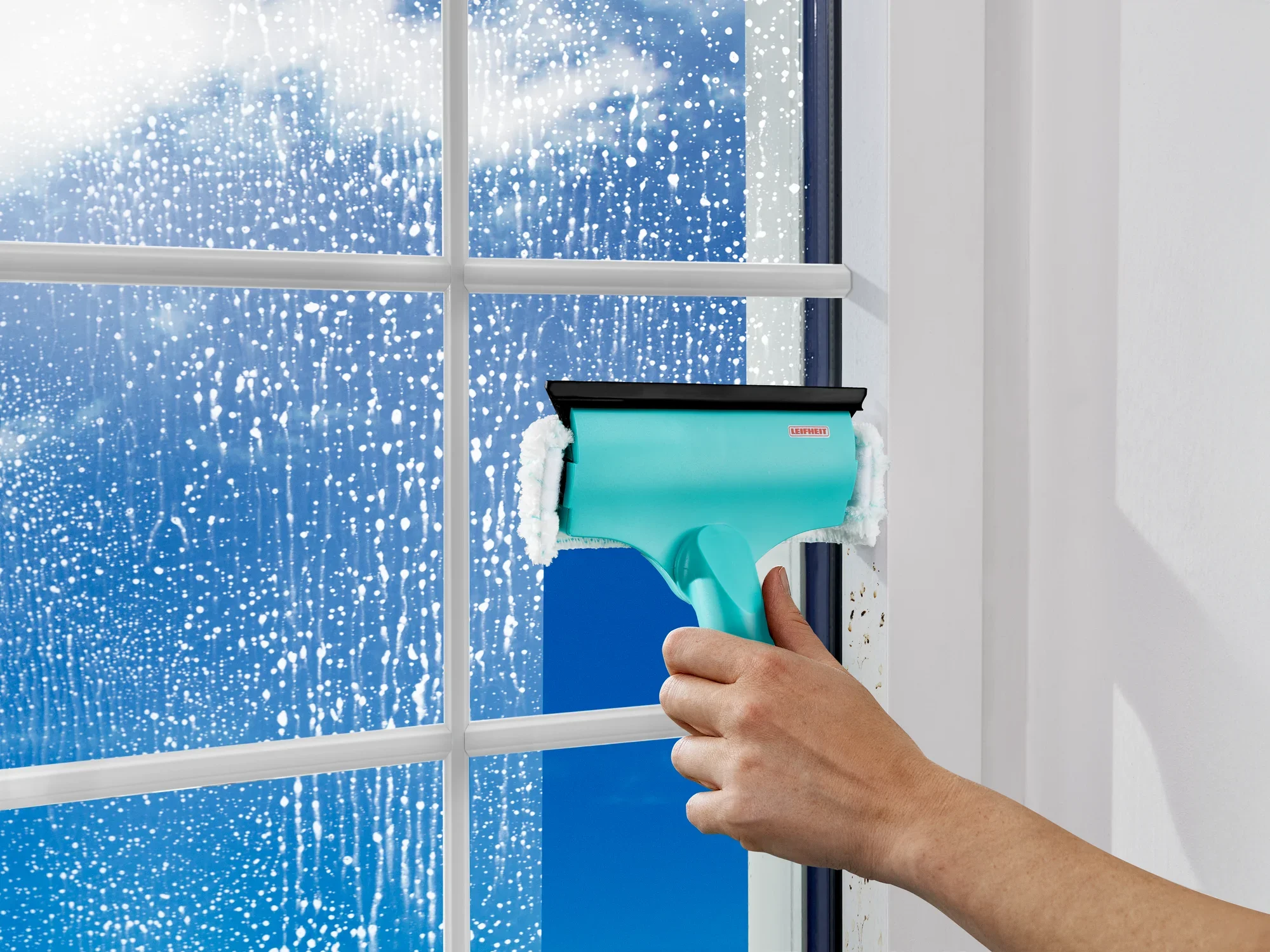 Mini ściągaczka do okien i ram 3 w 1 służy do czyszczenia okien. Ręka trzyma ściągaczkę w kolorze turkusowym, podczas gdy woda i piana są widoczne na szybie.
