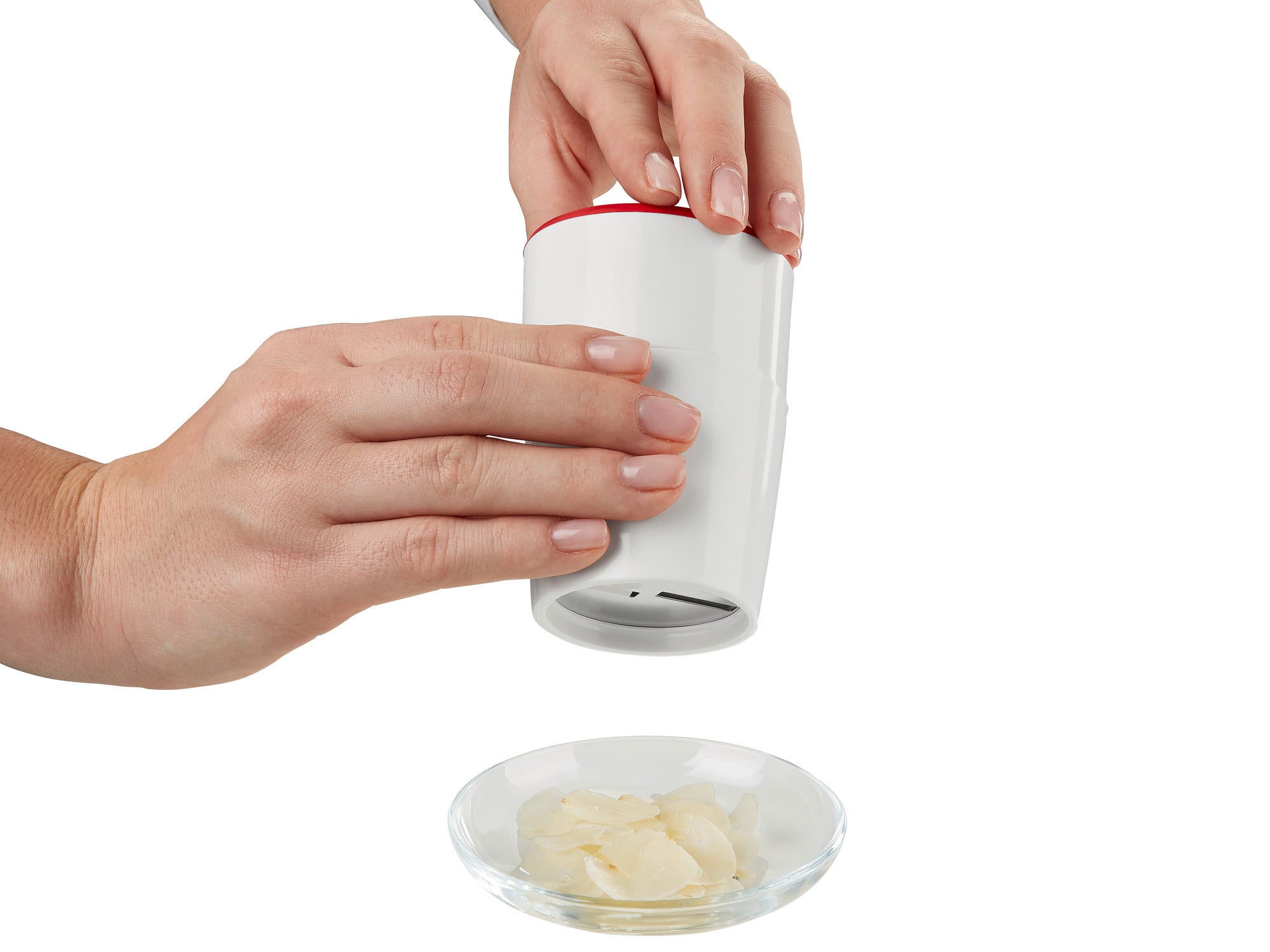 A garlic cutter is used to cut thin slices of garlic on a plate. Two hands hold the white cutter with red edge over the plate.