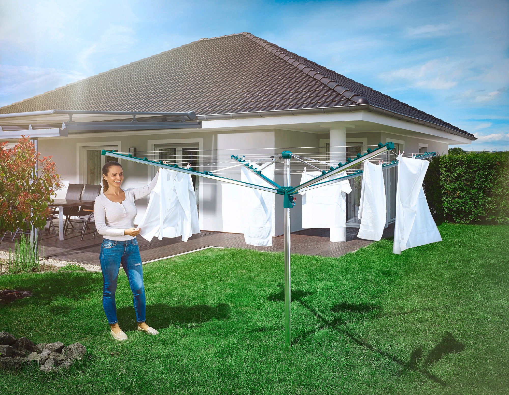 Een vrouw staat in de tuin voor een modern huis en gebruikt de Leifheit Linomatic 400 Comfort droogmolen. Witte was hangt aan de lijn te drogen.