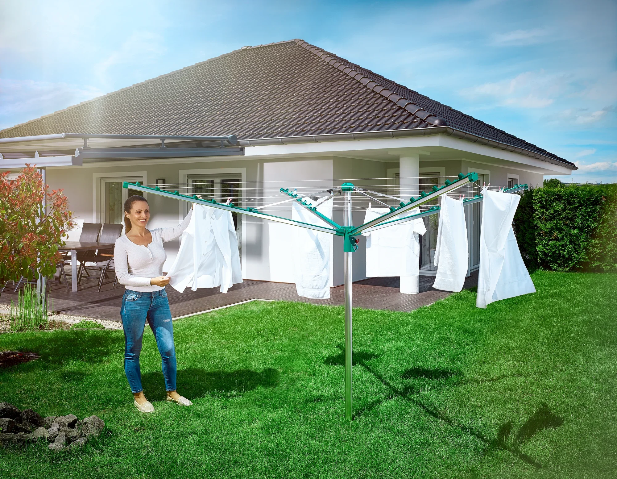 Une femme se tient dans le jardin devant une maison moderne et utilise le séchoir à linge Leifheit Linomatic 400 Comfort. Du linge blanc est suspendu sur les cordes pour sécher.