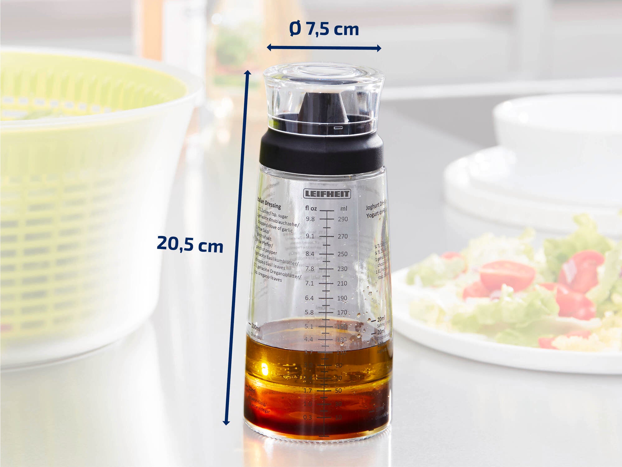 Ein transparenter Messbecher mit schwarzem Deckel der Marke Leifheit, gefüllt mit Öl. Der Becher ist 20,5 cm hoch und hat einen Durchmesser von 7,5 cm. Im Hintergrund ist eine Salatschüssel zu sehen.