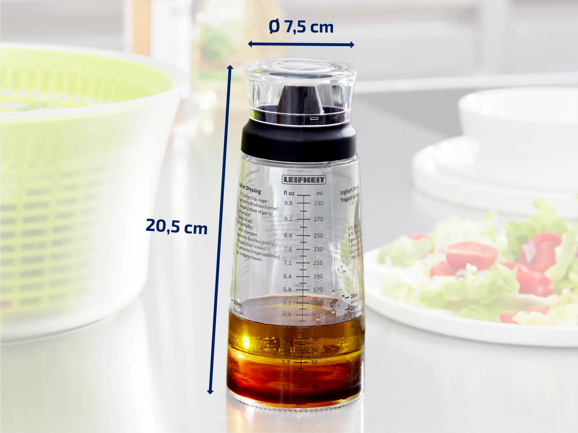 Een transparante maatbeker met een zwart deksel van het merk Leifheit, gevuld met olie. De beker is 20,5 cm hoog en heeft een diameter van 7,5 cm. Op de achtergrond is een slakom te zien.