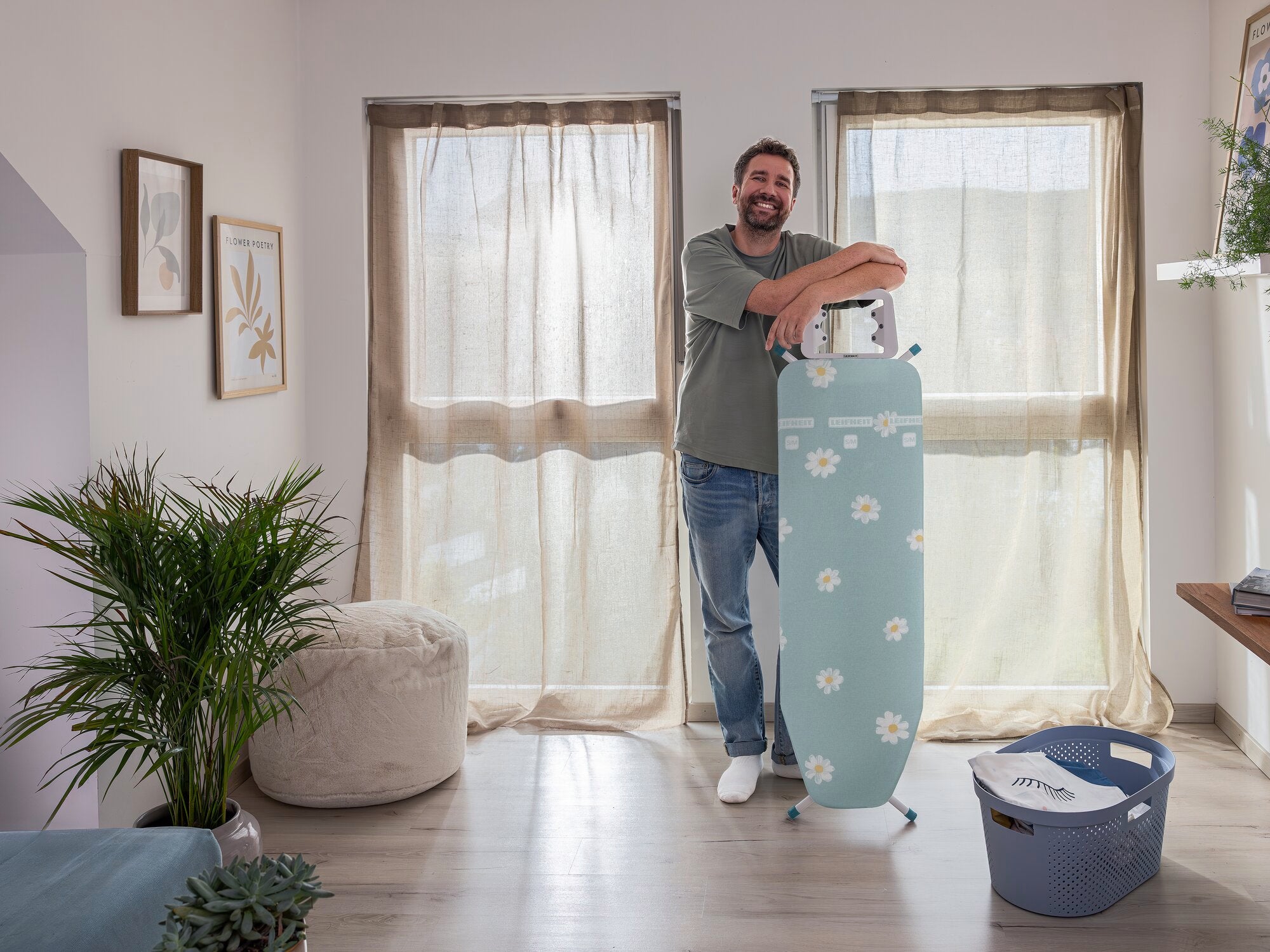 Ein Mann steht in einem hellen Raum neben einem Leifheit Classic M Basic Bügelbrett mit Blumenmuster. Im Hintergrund sind große Fenster mit Vorhängen und ein Wäschekorb zu sehen.