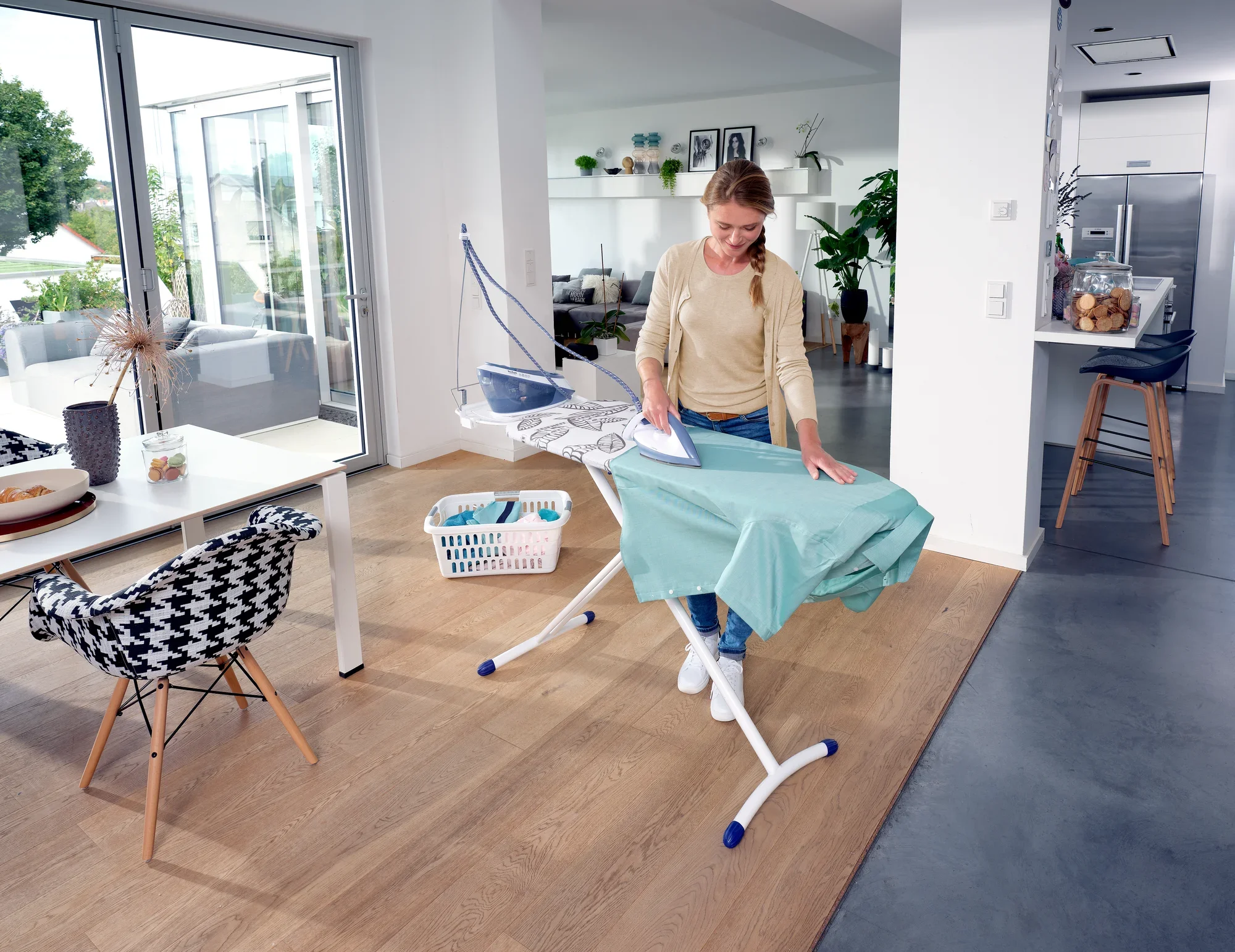 Une femme repasse une chemise sur la table à repasser Leifheit Air Board Express L Solid MAXX dans un salon moderne et lumineux. À l'arrière-plan, on aperçoit des plantes et une cuisine ouverte.