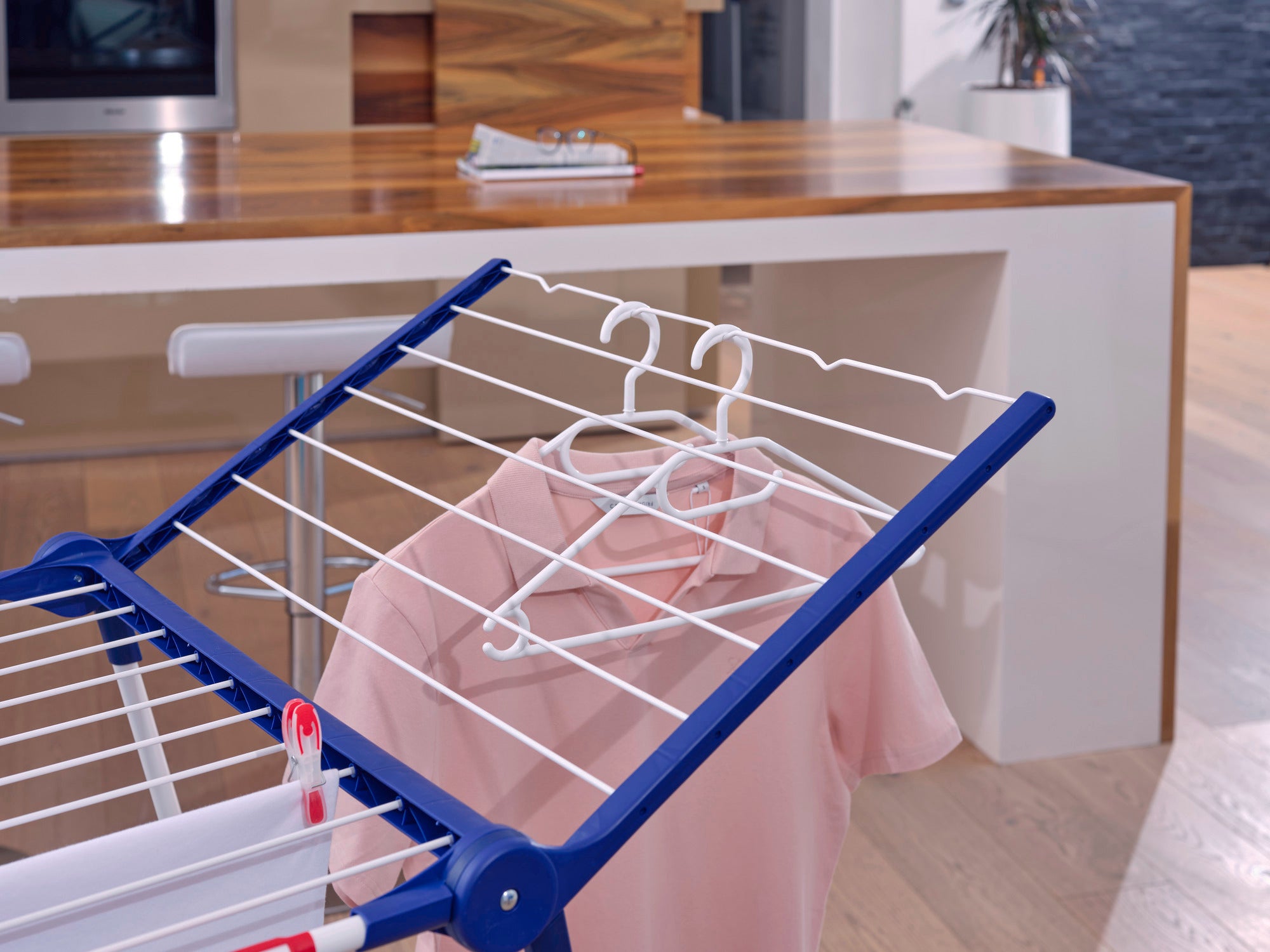 Een kledinghanger hangt aan een blauw droogrek in een moderne keuken. Een roze overhemd hangt aan de hanger. Op de achtergrond is een stijlvol keukenblok te zien.