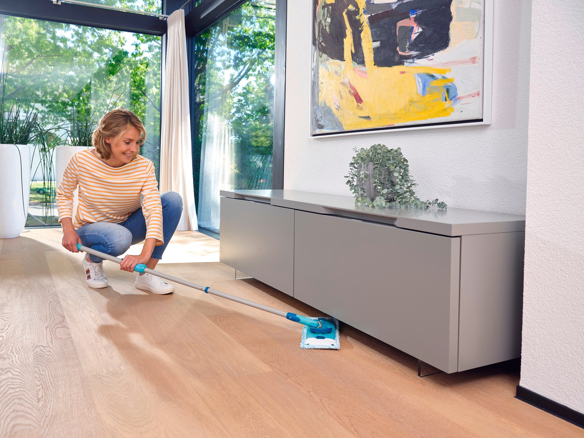 Een vrouw reinigt de houten vloer van een moderne woonkamer met de CLEAN TWIST M Ergo. Op de achtergrond zijn grote ramen en een abstract schilderij te zien.