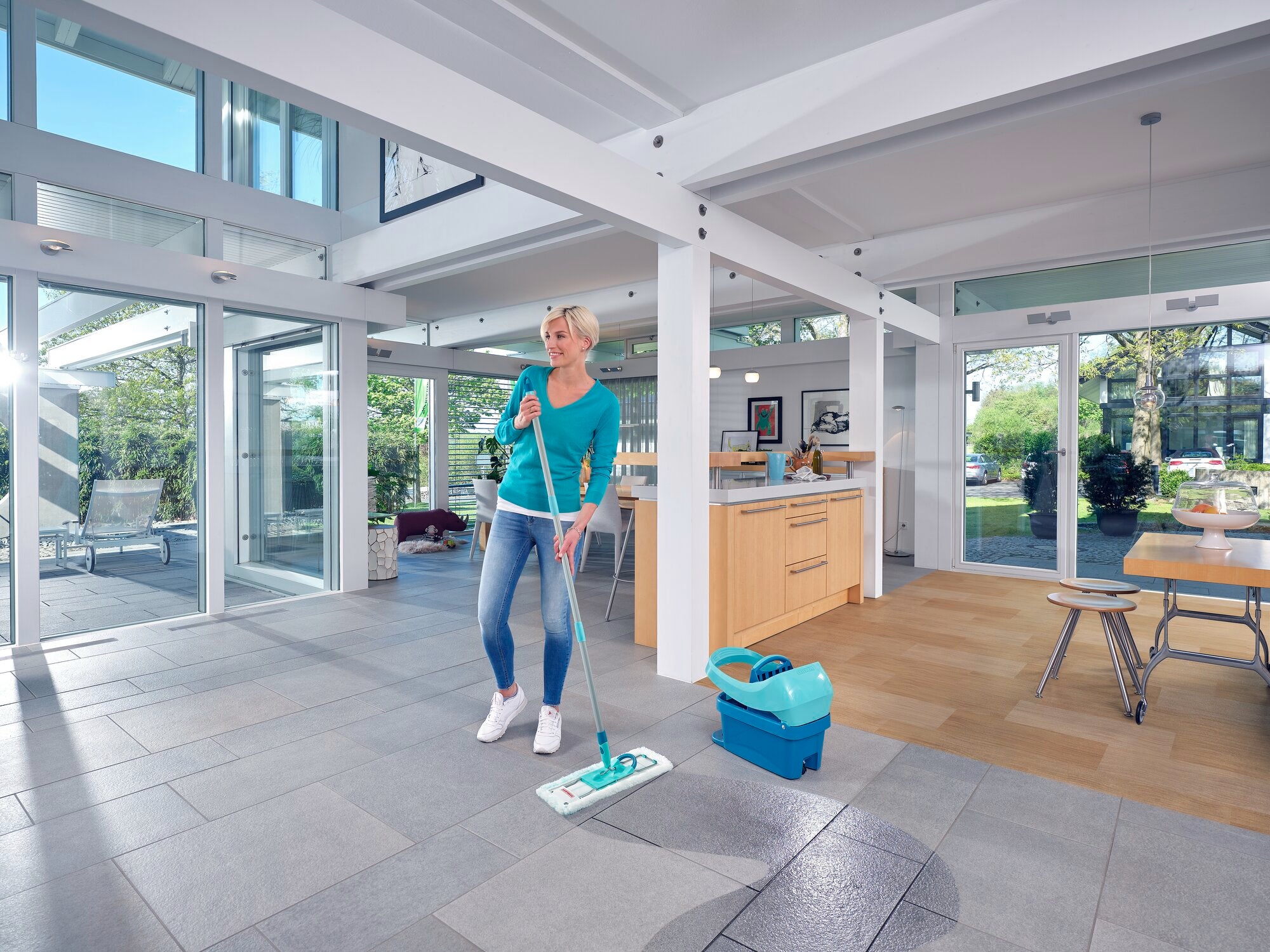 Una mujer limpia el suelo en una habitación moderna y luminosa con la prensa para paños Profi XL. La habitación tiene grandes ventanas y una cocina abierta, lo que crea un ambiente luminoso y acogedor.