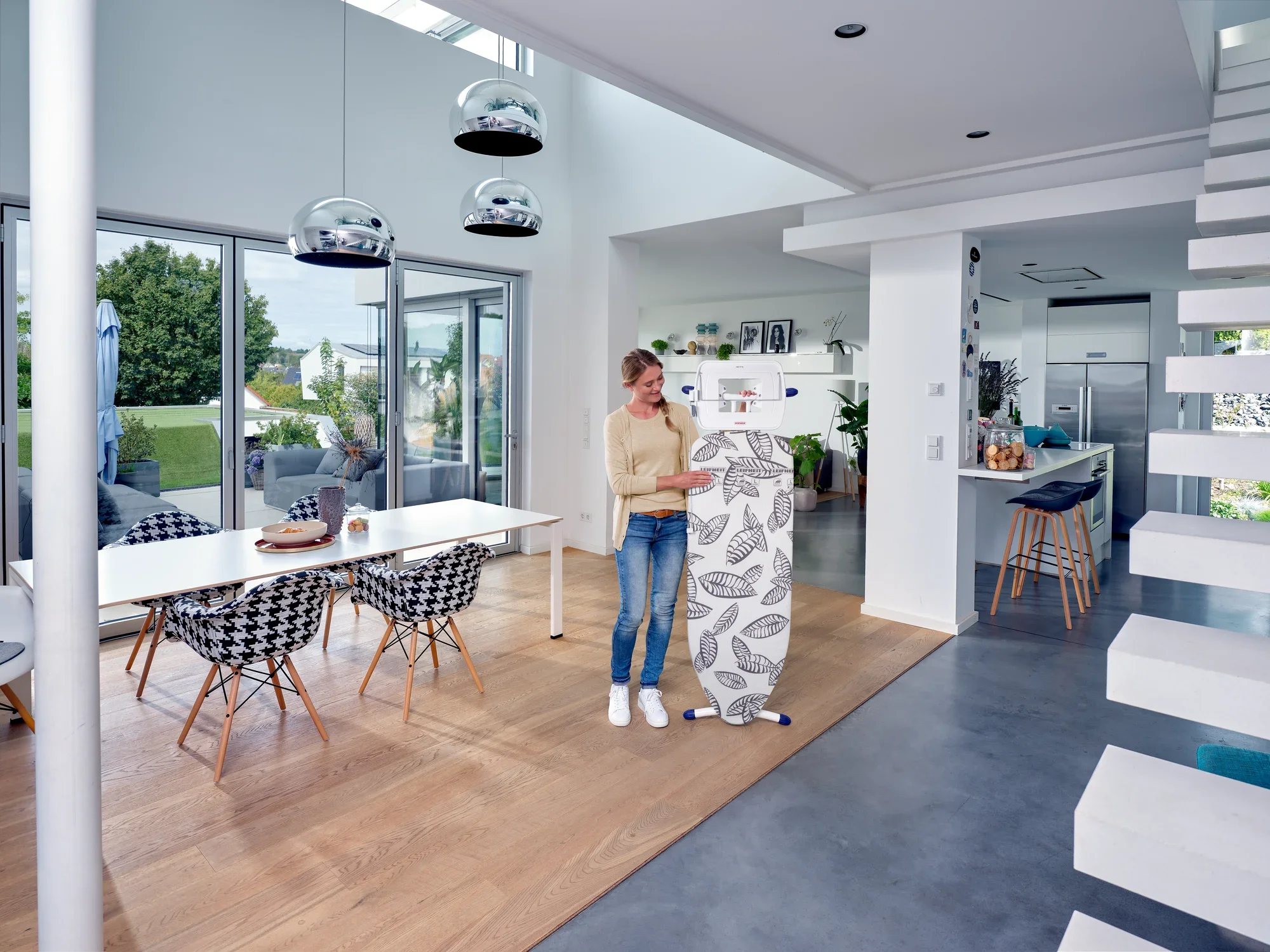 Une femme se tient dans un salon moderne et lumineux et utilise la table à repasser Air Board Express L Solid MAXX. La pièce est meublée avec style, avec une grande table et des chaises design.