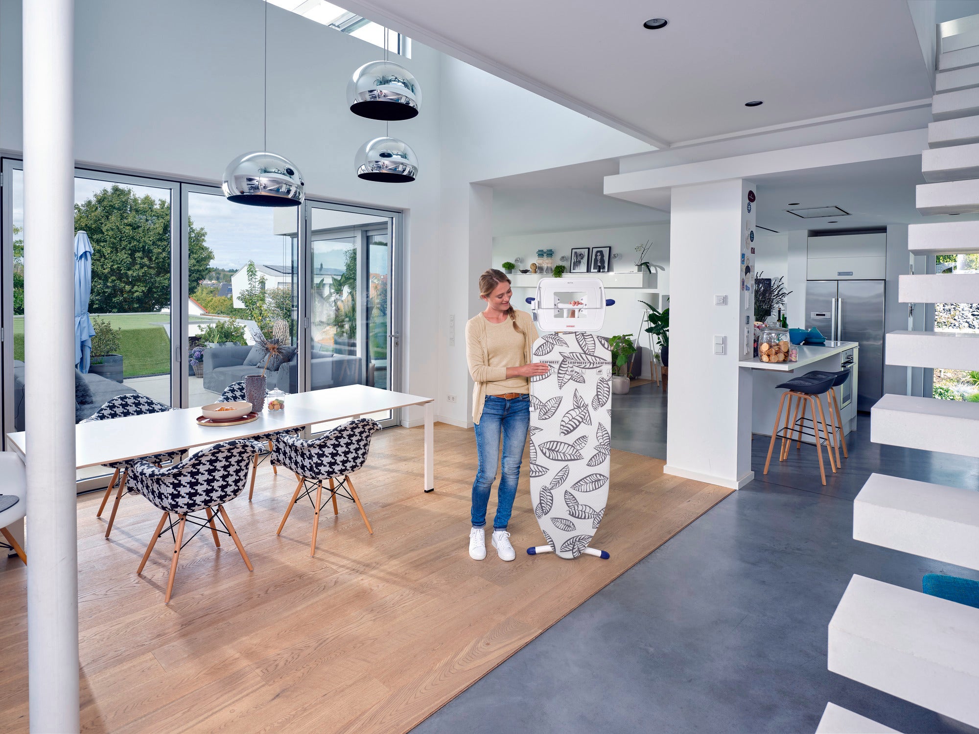 Une femme se tient dans un salon moderne et lumineux et utilise la table à repasser Air Board Express L Solid MAXX. La pièce est meublée avec style, avec une grande table et des chaises design.