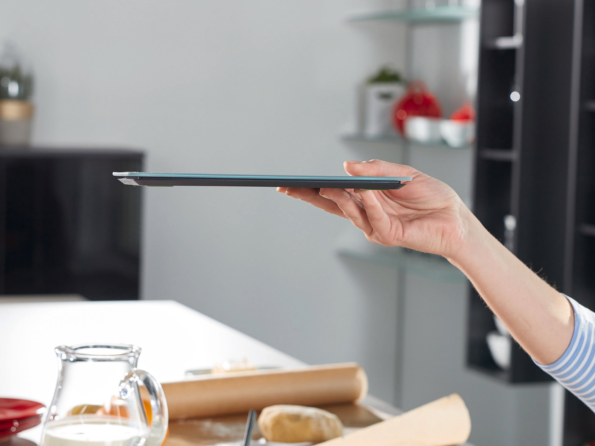 Jedna ręka trzyma cyfrową wagę kuchenną Page Comfort 300 Slim, która charakteryzuje się smukłą konstrukcją. W tle widoczne są przybory kuchenne.