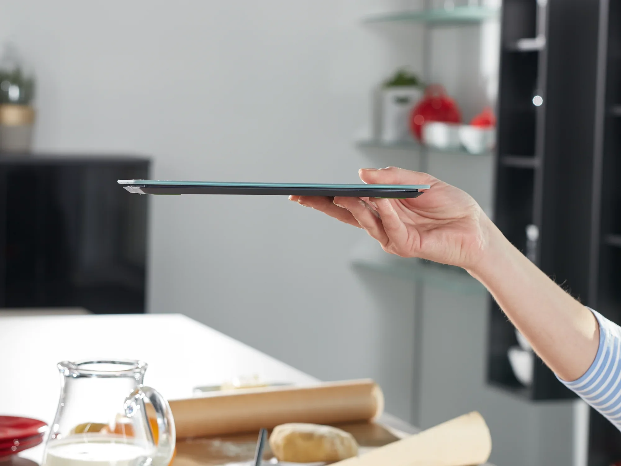 Eén hand houdt de Page Comfort 300 Slim digitale keukenweegschaal vast, die wordt gekenmerkt door zijn slanke ontwerp. Op de achtergrond is keukengerei te zien.