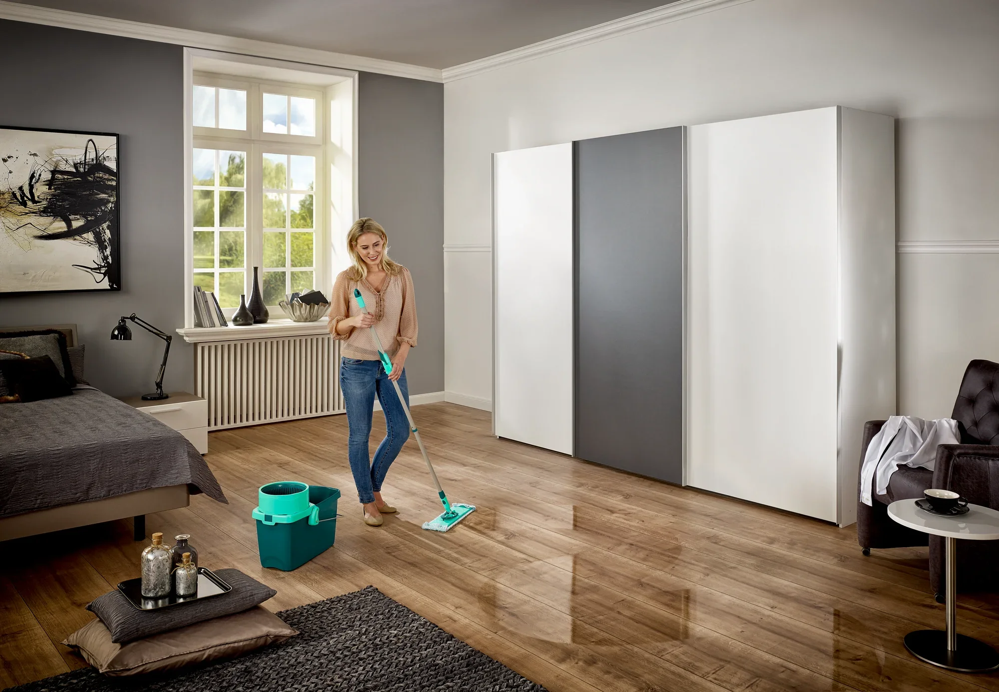 Eine Frau reinigt den Holzboden eines modernen Schlafzimmers mit dem CLEAN TWIST System M. Neben ihr steht ein Eimer, der Teil des Sets ist. Das Zimmer ist hell und stilvoll eingerichtet.