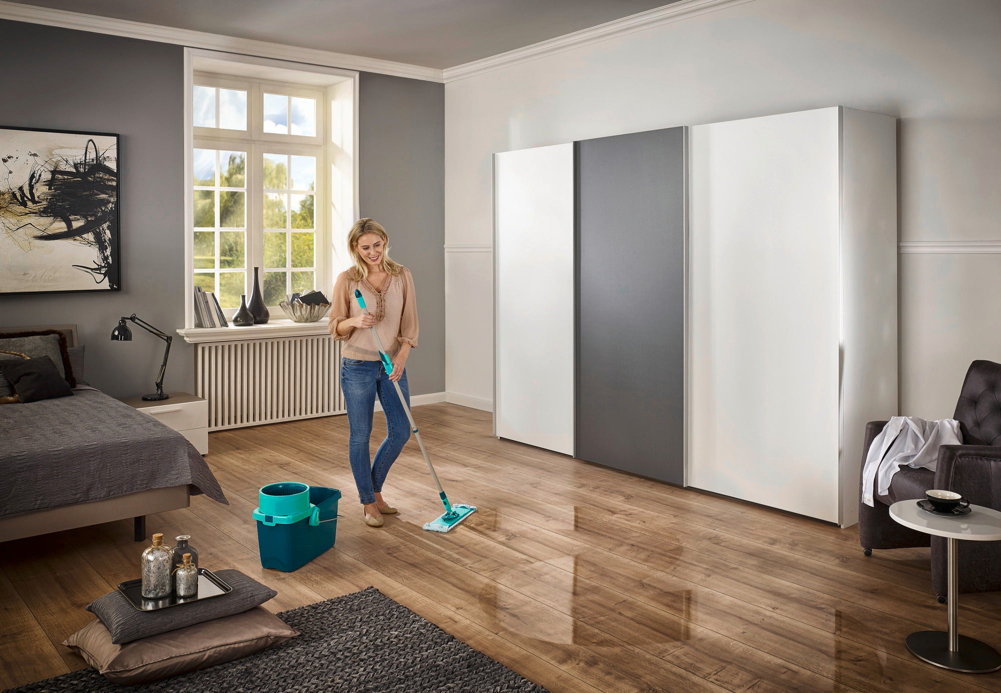 Eine Frau reinigt den Holzboden eines modernen Schlafzimmers mit dem CLEAN TWIST System M. Neben ihr steht ein Eimer, der Teil des Sets ist. Das Zimmer ist hell und stilvoll eingerichtet.