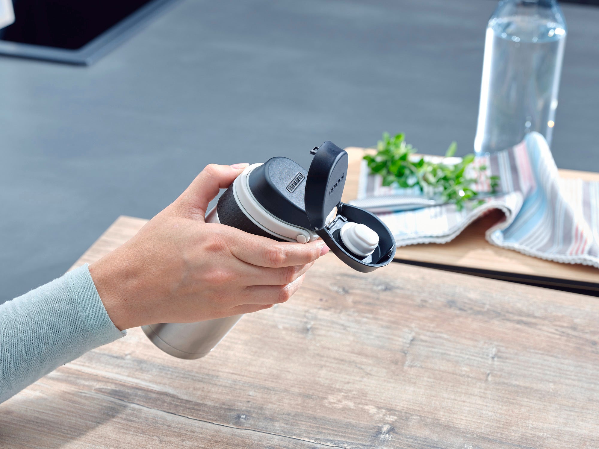 A person is holding a silver Flip insulated mug with a capacity of 350 ml. The lid is open and a towel and a water bottle can be seen in the background.