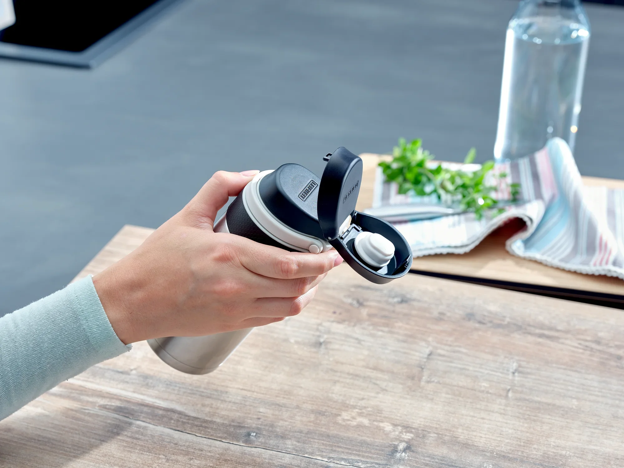 Osoba trzyma srebrny izolowany kubek Flip o pojemności 350 ml. Pokrywka jest otwarta, a w tle widać ręcznik i butelkę wody.