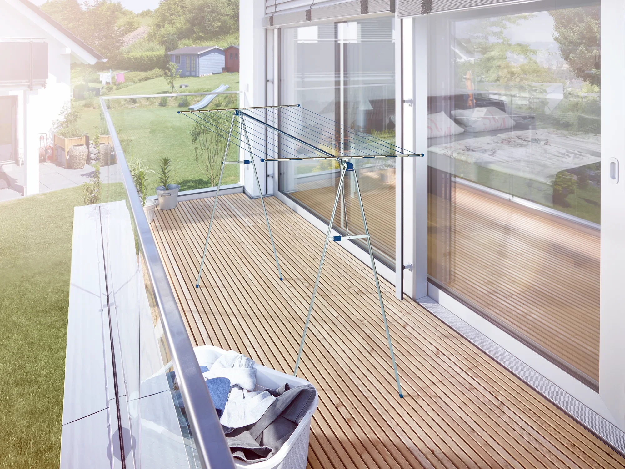 Wysoka suszarka Linomaxx 210 stoi na nowoczesnym balkonie z drewnianymi deskami. W tle widać duże okna i zielony krajobraz. Obok stoi kosz na pranie.