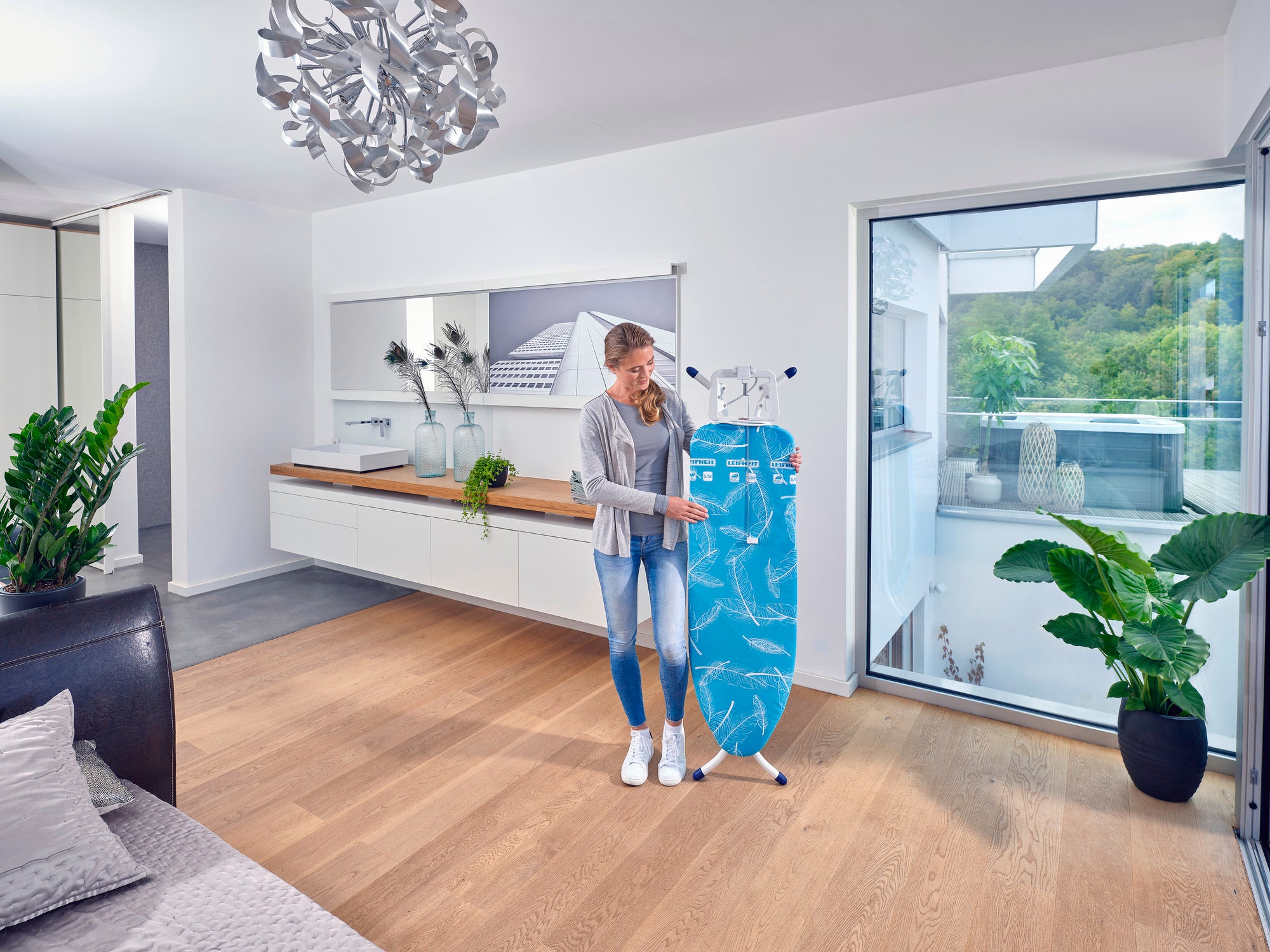 Eine Frau steht in einem modernen, hellen Raum und hält einen Bügeltisch namens Air Board M Compact Plus. Der Raum hat Holzböden, große Fenster und stilvolle Dekorationen.