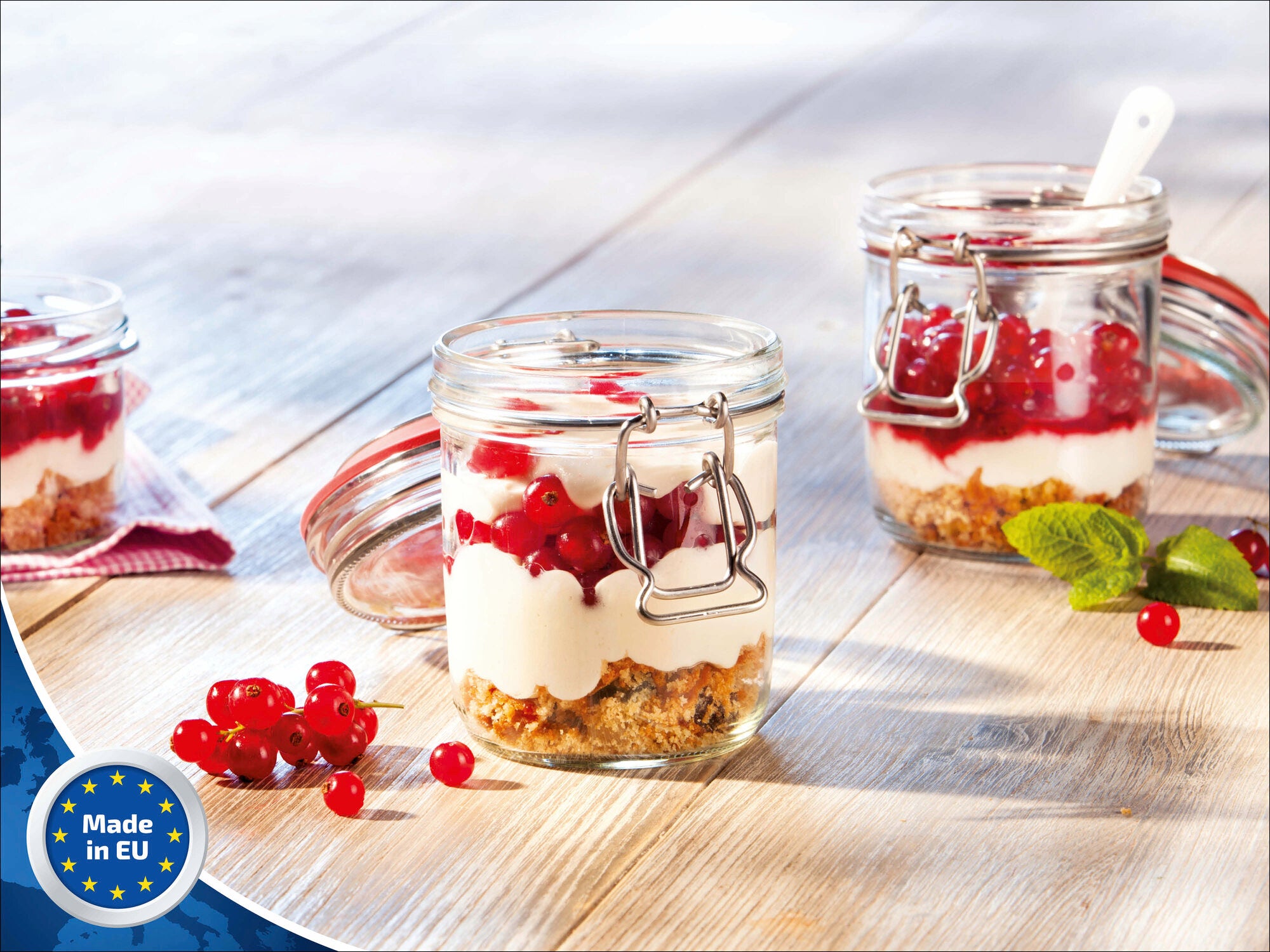 Drei Leifheit Drahtbügelgläser mit 370 ml Fassungsvermögen stehen auf einem Holztisch. Sie sind mit Joghurt, roten Johannisbeeren und Müsli gefüllt. Ein Glas ist geöffnet, daneben liegen frische Beeren.