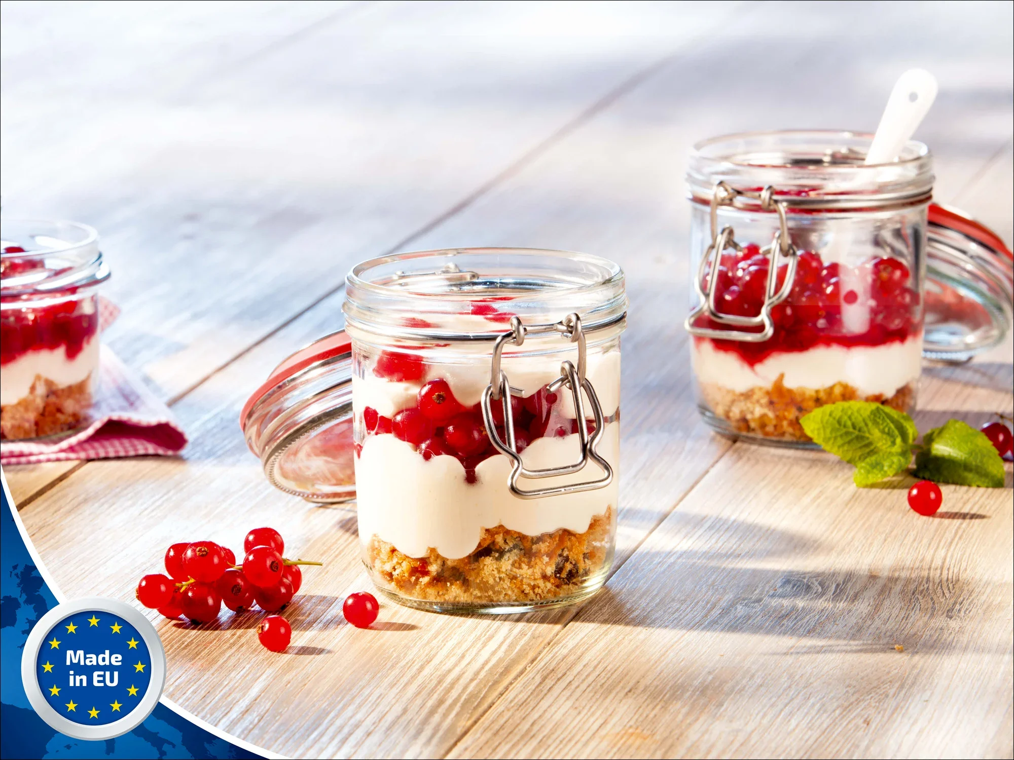 Drei Leifheit Drahtbügelgläser mit 370 ml Fassungsvermögen stehen auf einem Holztisch. Sie sind mit Joghurt, roten Johannisbeeren und Müsli gefüllt. Ein Glas ist geöffnet, daneben liegen frische Beeren.