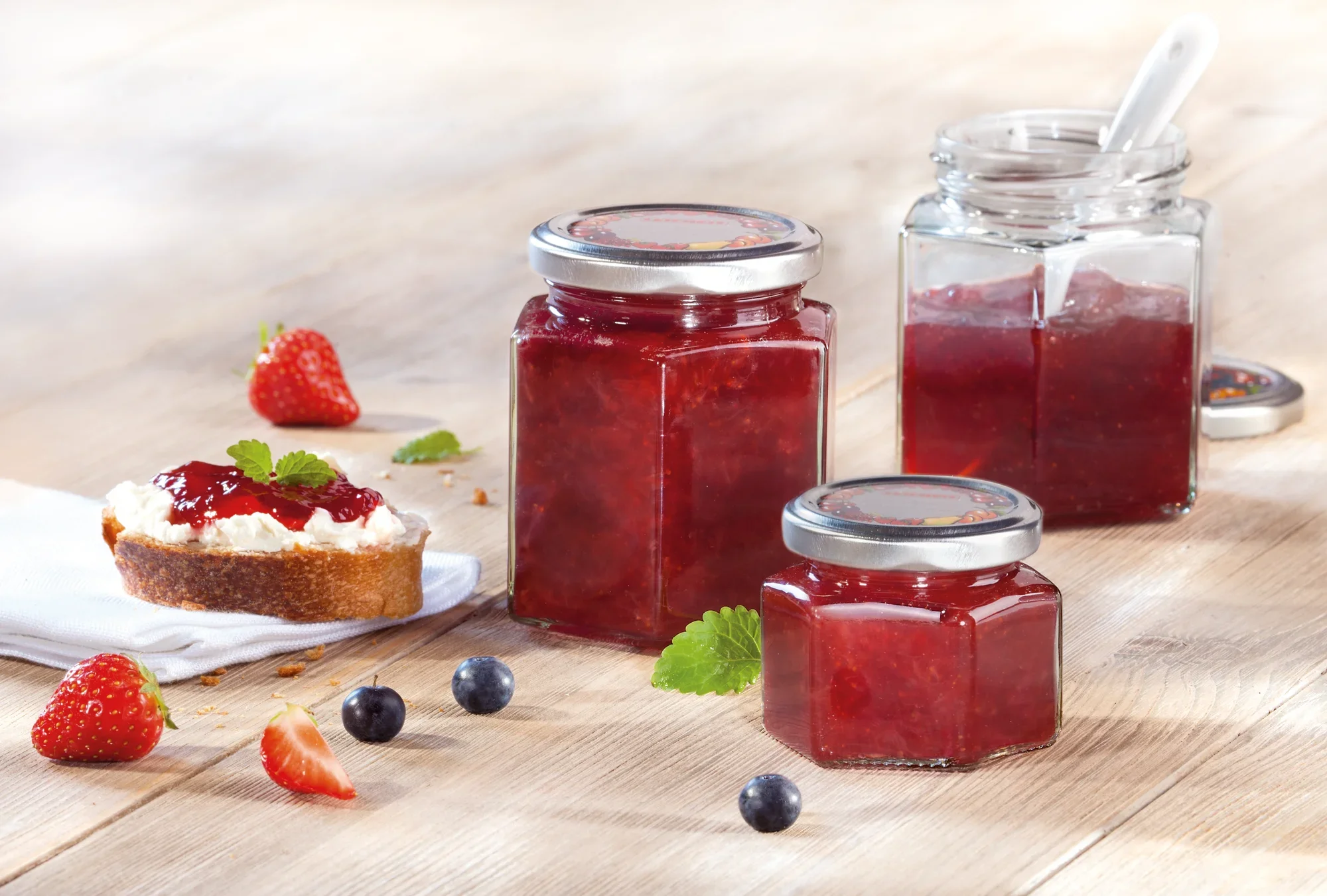 Trzy sześciokątne słoiki z czerwonym dżemem na drewnianym stole. Obok znajduje się kromka chleba z dżemem, udekorowana truskawkami i listkami mięty. W tle świeże jagody.