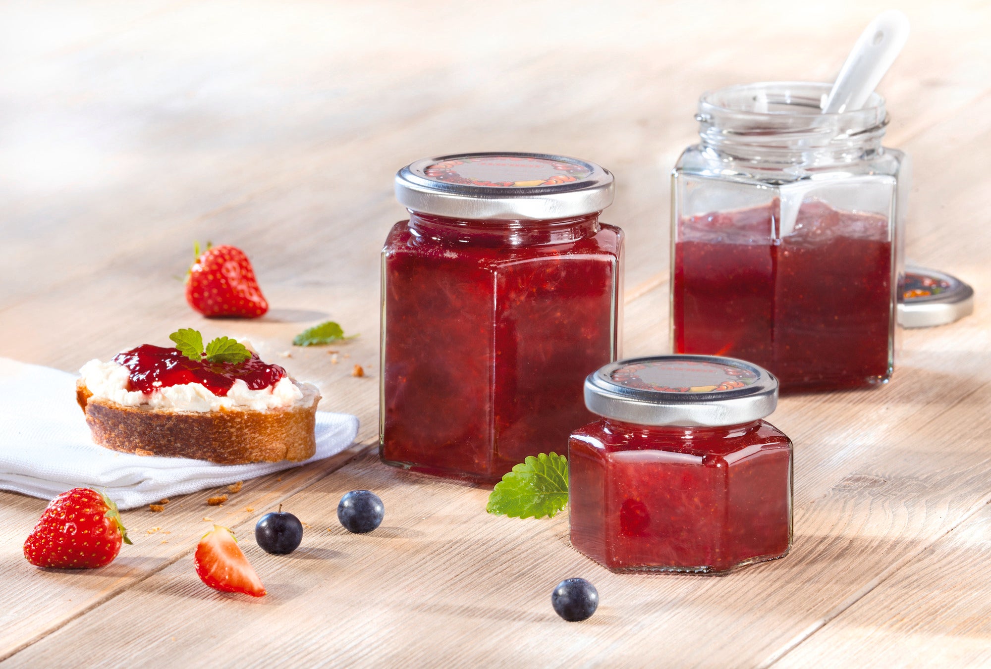 Trois bocaux hexagonaux contenant de la confiture rouge sur une table en bois. À côté, une tranche de pain tartinée de confiture, garnie de fraises et de feuilles de menthe. Des baies fraîches sont disposées en arrière-plan.