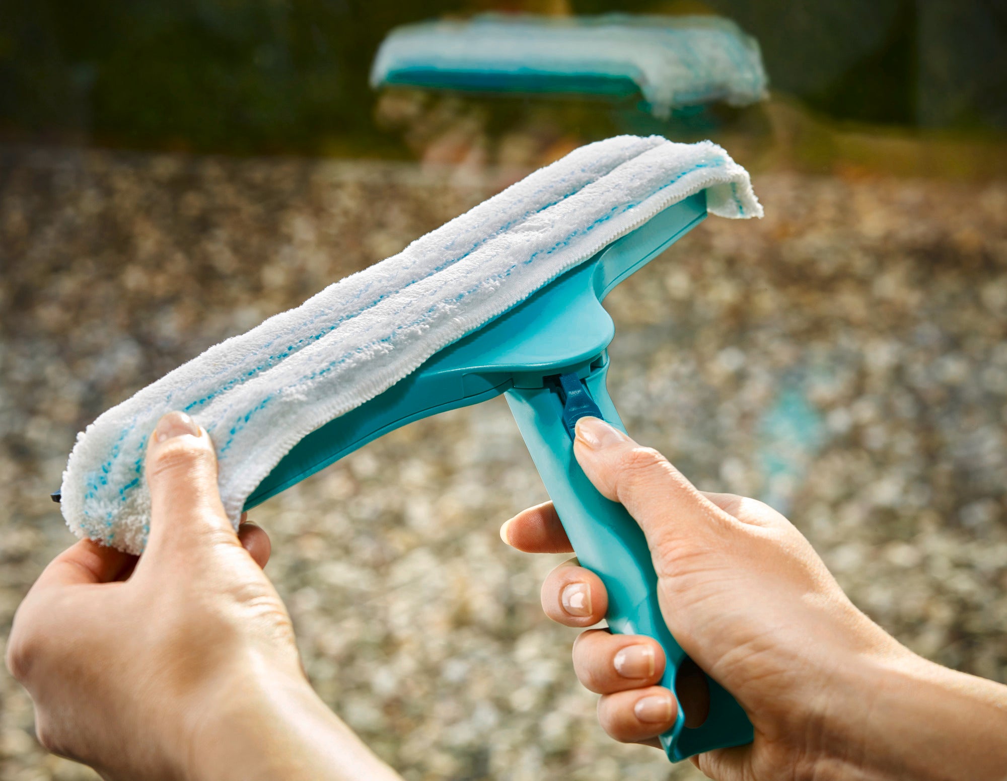 Raamwisser 3in1 in gebruik. Eén persoon houdt de turquoise raamwisser met een witte microvezelhoes vast terwijl hij een raam schoonmaakt. Ideaal voor streeploos schoonmaken.