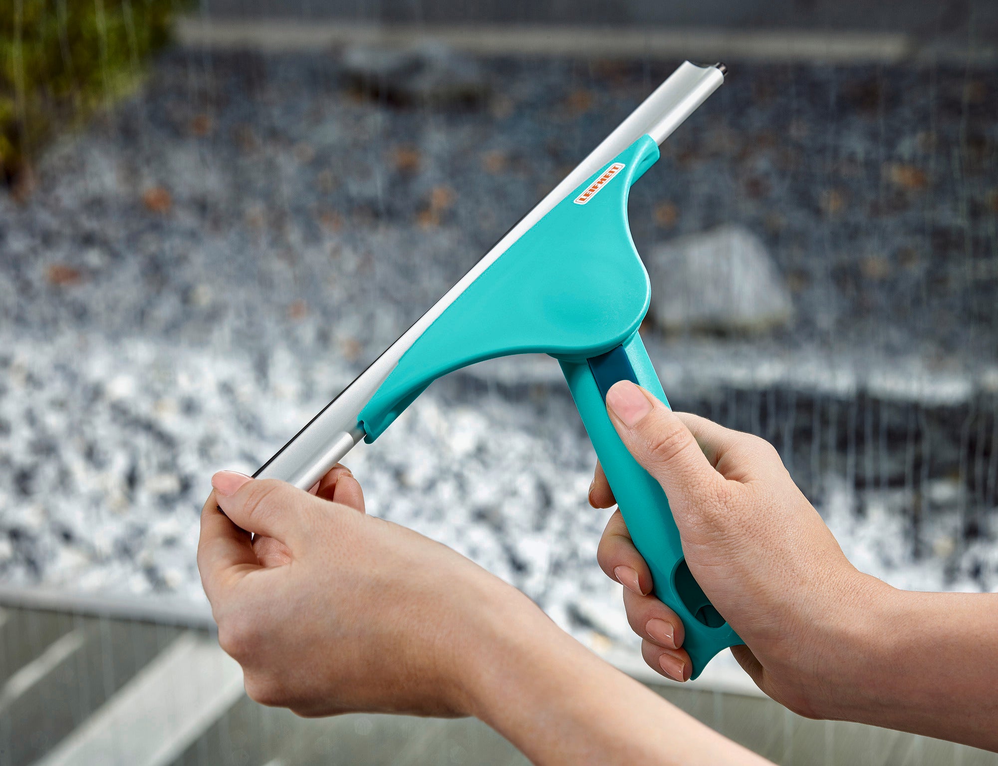 Fensterabzieher Window Slider M in Aktion. Eine Person hält den türkisfarbenen Abzieher mit ergonomischem Griff, um Wasser von einer Glasoberfläche zu entfernen. Ideal für streifenfreie Reinigung.