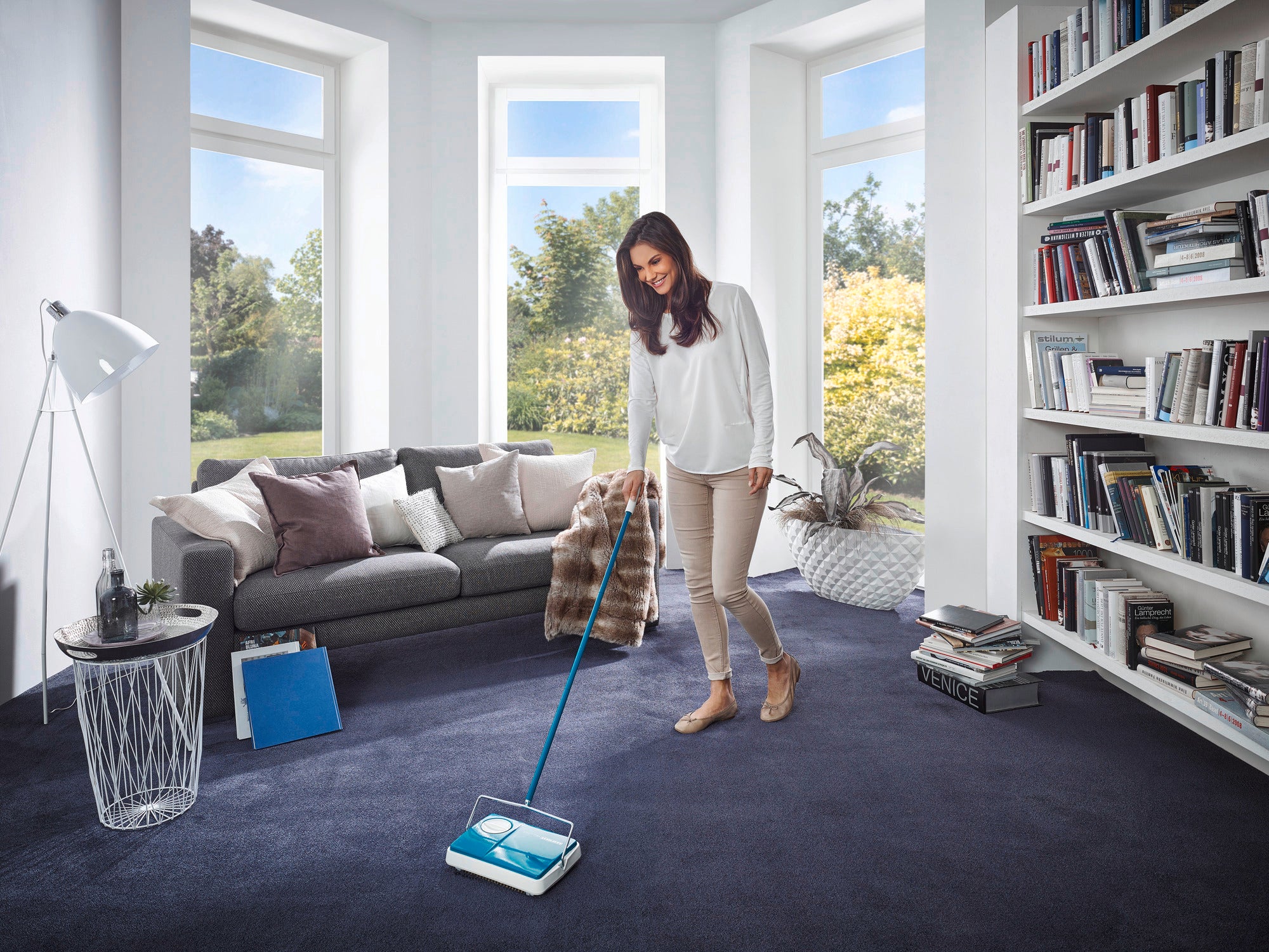A woman uses the turquoise Regulus carpet sweeper in a bright living room with large windows. The room is modernly furnished with a grey sofa and a bookcase.