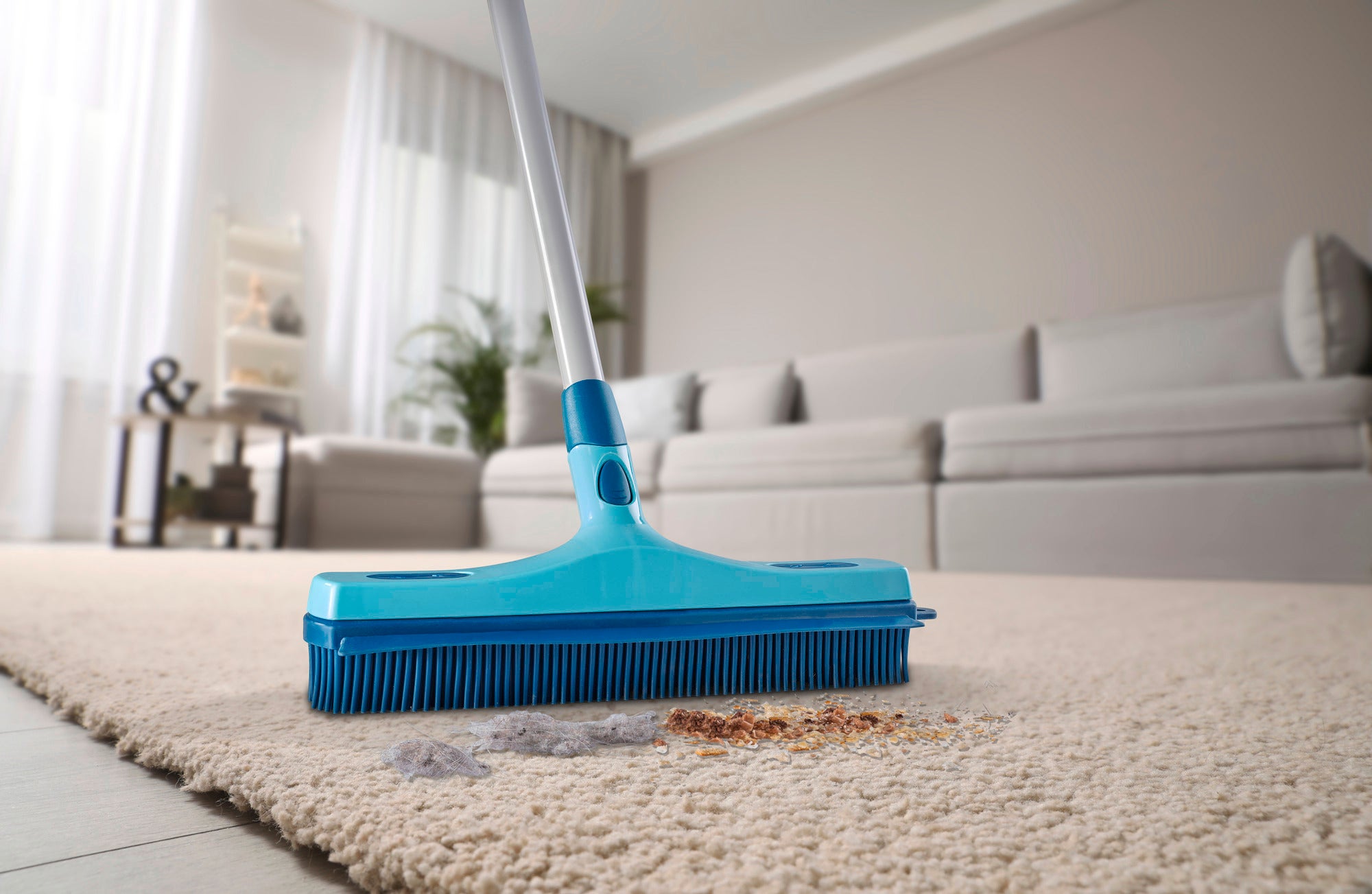 A blue rubber brush, model Profi XL, is used to remove dirt from a light-coloured carpet in a modern living room. A sofa and plants can be seen in the background.