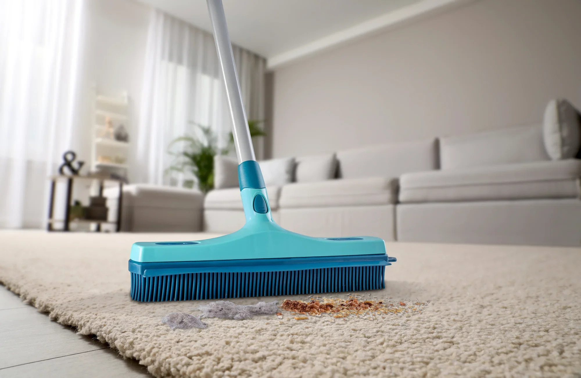 Een blauwe rubberen bezem, model Profi XL, wordt gebruikt om vuil te verwijderen van een lichtgekleurd tapijt in een moderne woonkamer. Op de achtergrond zijn een bank en planten te zien.