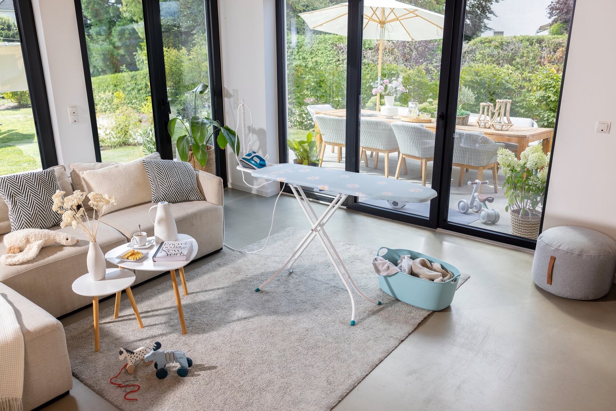 Bügelbrett Leifheit Classic Basic Plus in einem modernen Wohnzimmer. Es steht auf einem Teppich, umgeben von einem Sofa, Couchtischen und Spielzeug. Große Fenster bieten Blick auf einen Garten.