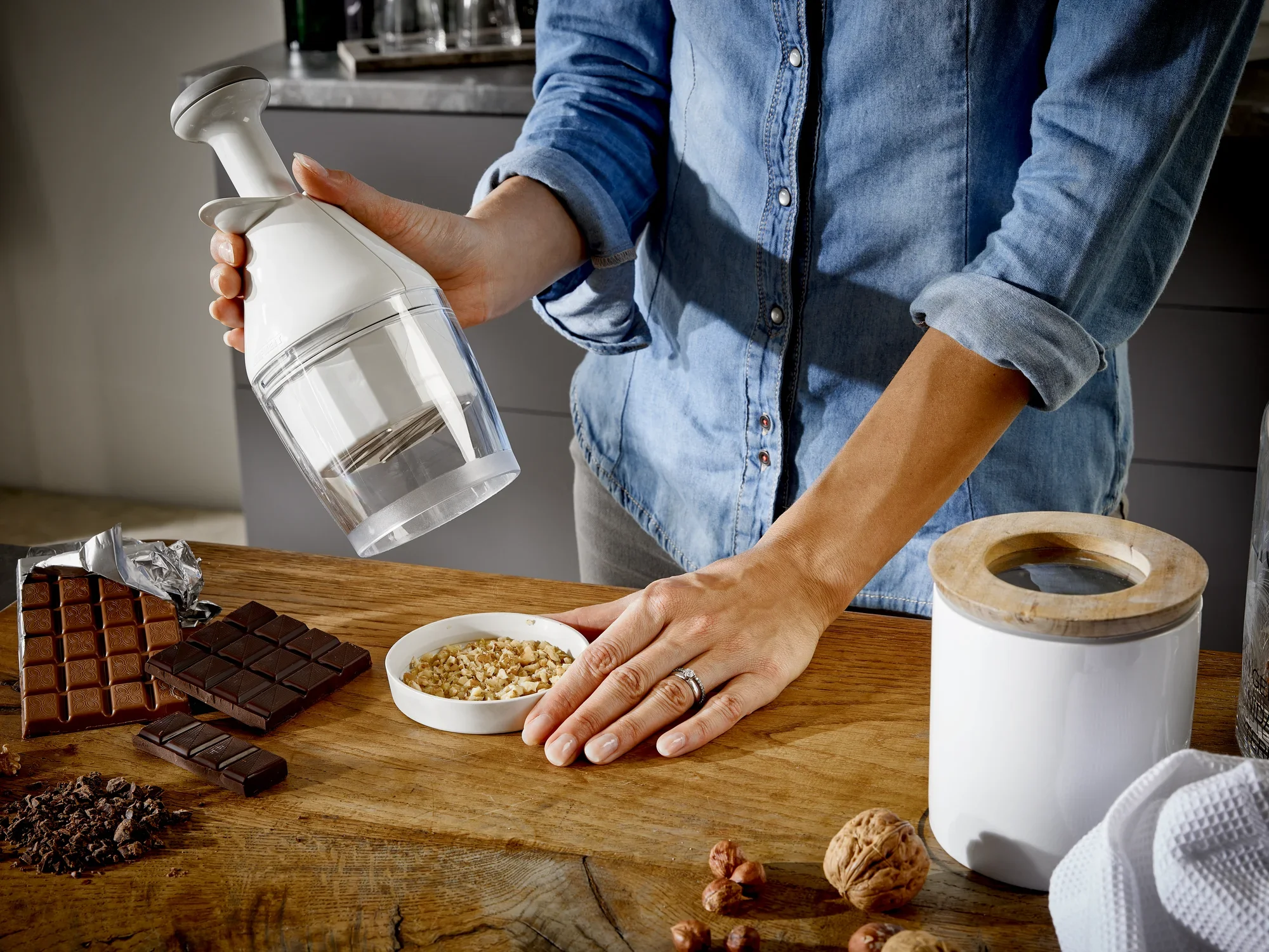 Zwiebel- und Gemüsehacker in Anwendung: Eine Person hackt Nüsse mit einem weißen Zwiebel- und Gemüsehacker auf einem Holztisch. Daneben liegen Schokolade und ein Vorratsbehälter.