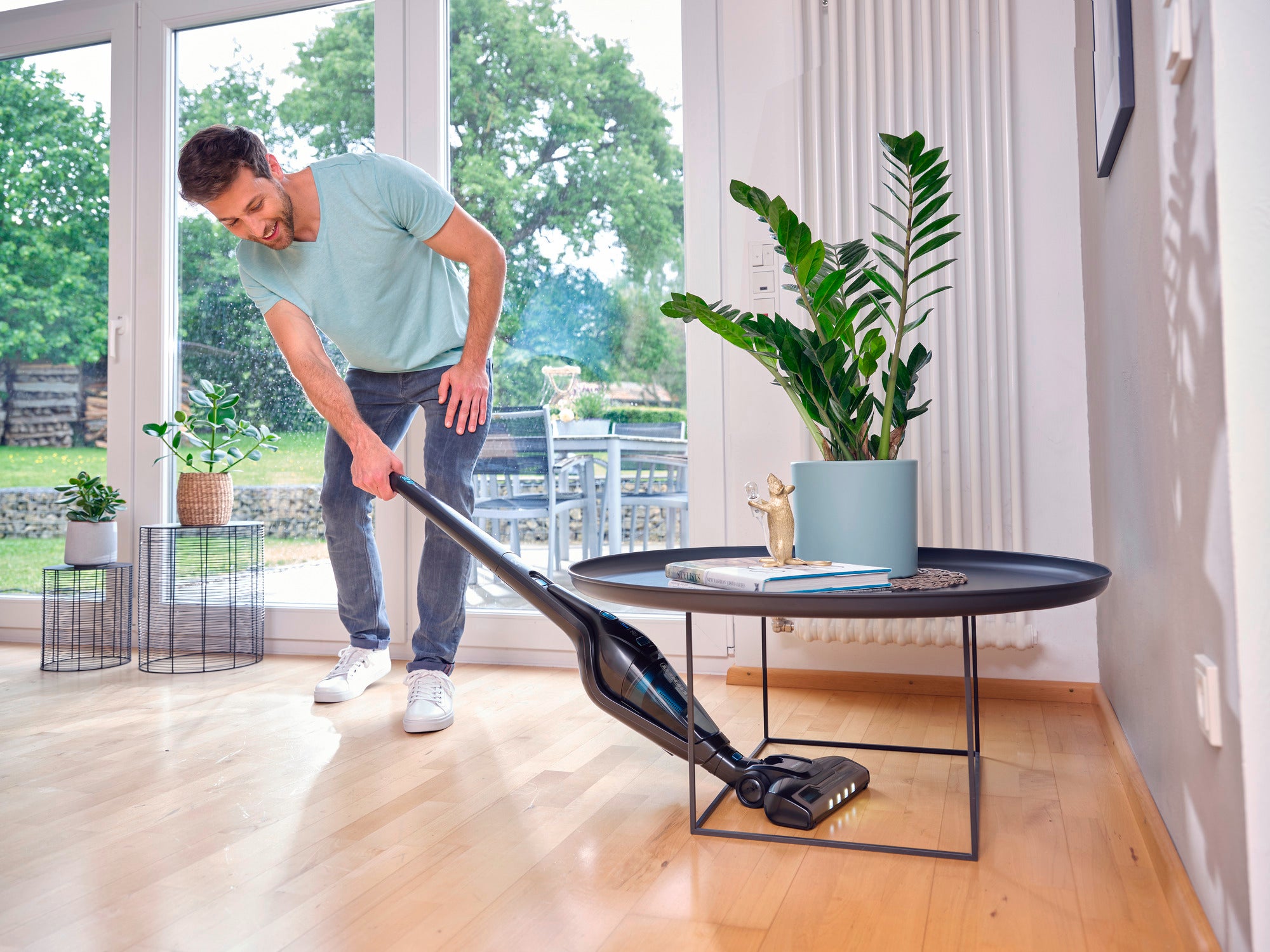 Ein Mann benutzt den Akku-Staubsauger Rotaro PowerVac, um den Holzboden in einem modernen Wohnzimmer zu reinigen. Im Hintergrund sind große Fenster und Pflanzen zu sehen.