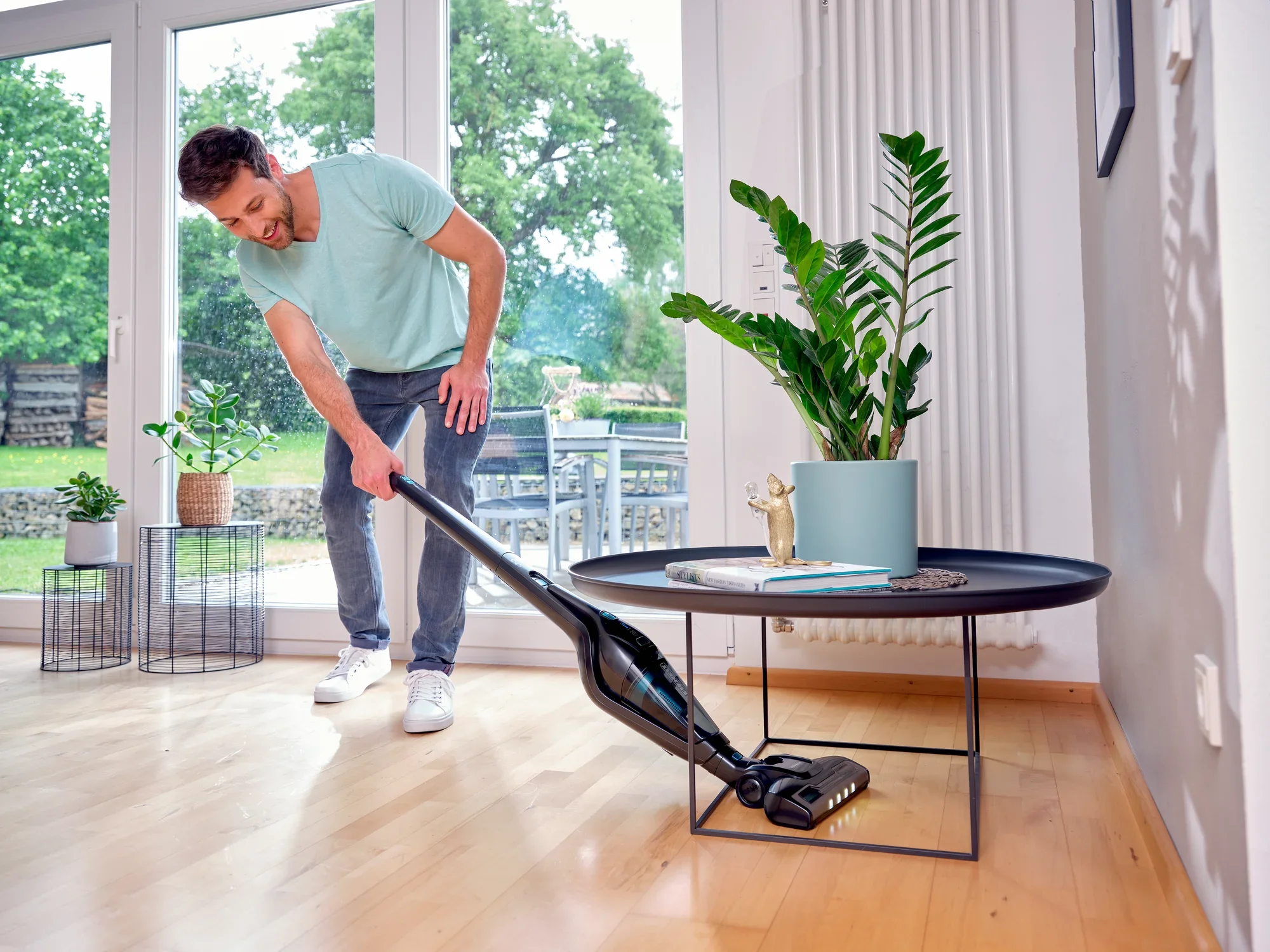 Un homme utilise l'aspirateur sans fil Rotaro PowerVac pour nettoyer le parquet d'un salon moderne. On aperçoit de grandes fenêtres et des plantes à l'arrière-plan.