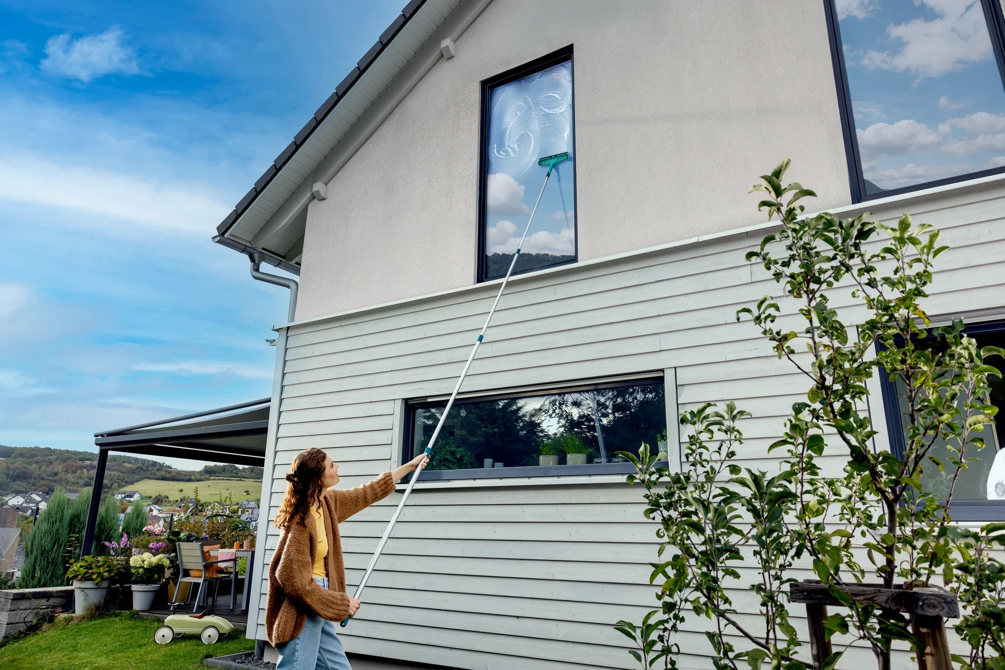 Jedna osoba czyści wysokie okno domu za pomocą trzonka teleskopowego, który można wydłużyć z 145 do 400 cm. Uchwyt umożliwia dotarcie do trudno dostępnych miejsc.