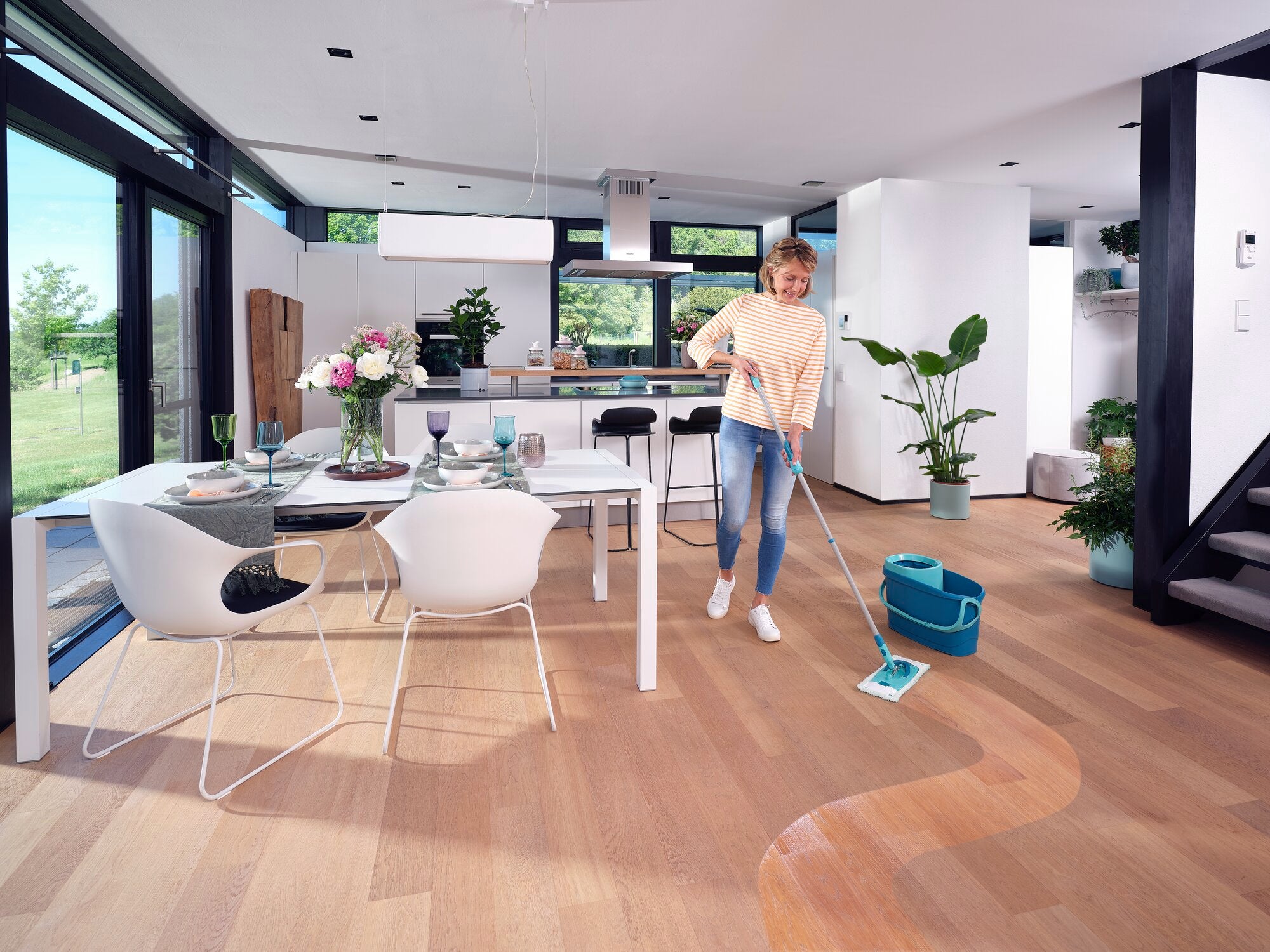 Eine Frau reinigt den Holzboden eines modernen, lichtdurchfluteten Wohnzimmers mit dem CLEAN TWIST M Ergo. Der Raum ist stilvoll eingerichtet mit einem Esstisch, Stühlen und Pflanzen.
