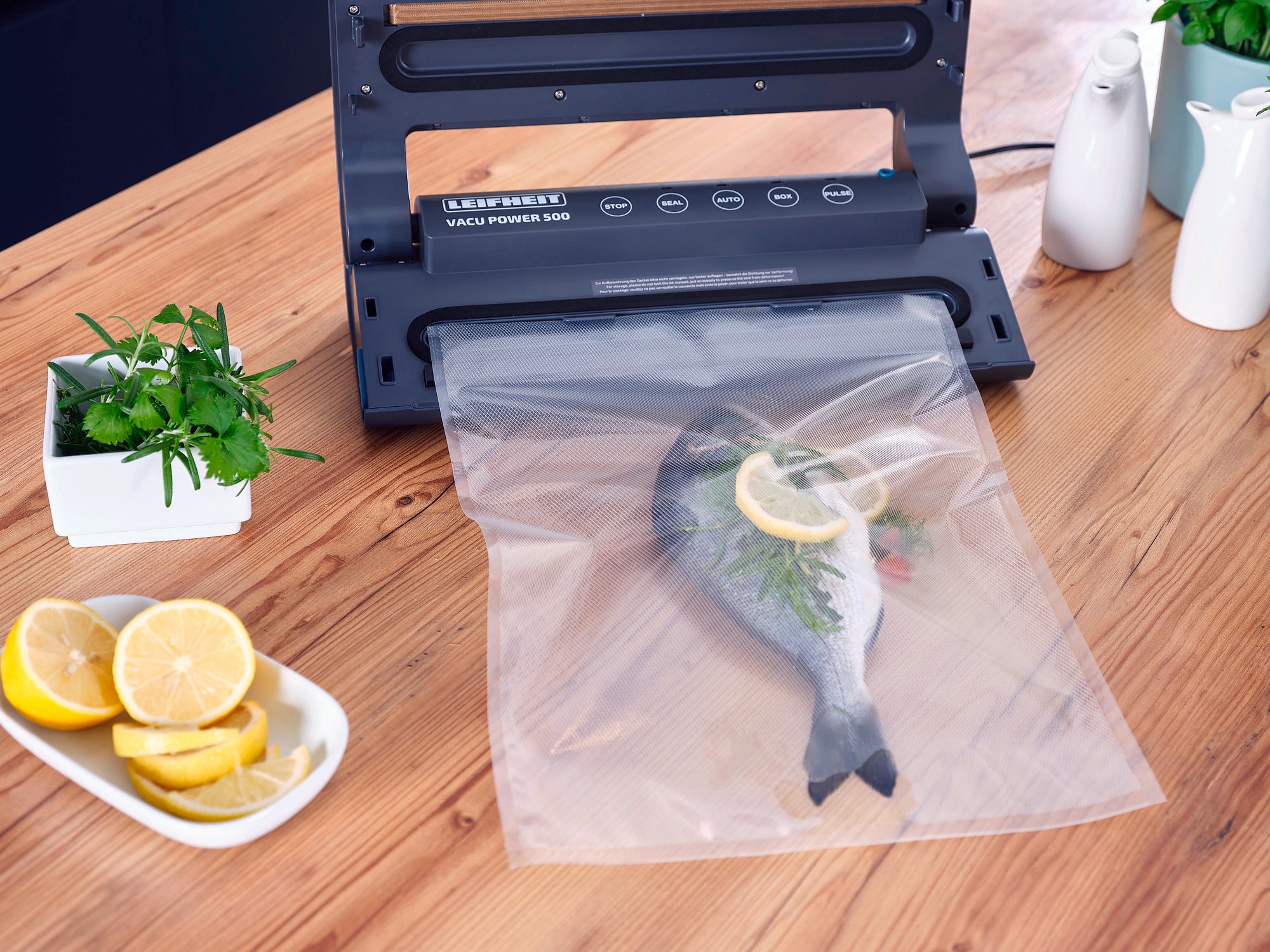 Sacchetti sottovuoto 30x40 cm: una macchina sottovuoto chiude un sacchetto con un pesce, guarnito con una fetta di limone ed erbe aromatiche. Accanto ci sono fette di limone ed erbe aromatiche fresche.