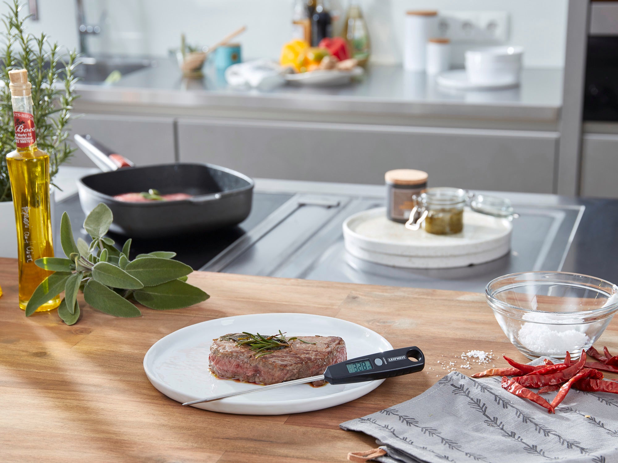 Een digitale universele thermometer ligt op een bord met een biefstuk in een moderne keuken. Kookgerei en kruiden zijn te zien op de achtergrond.