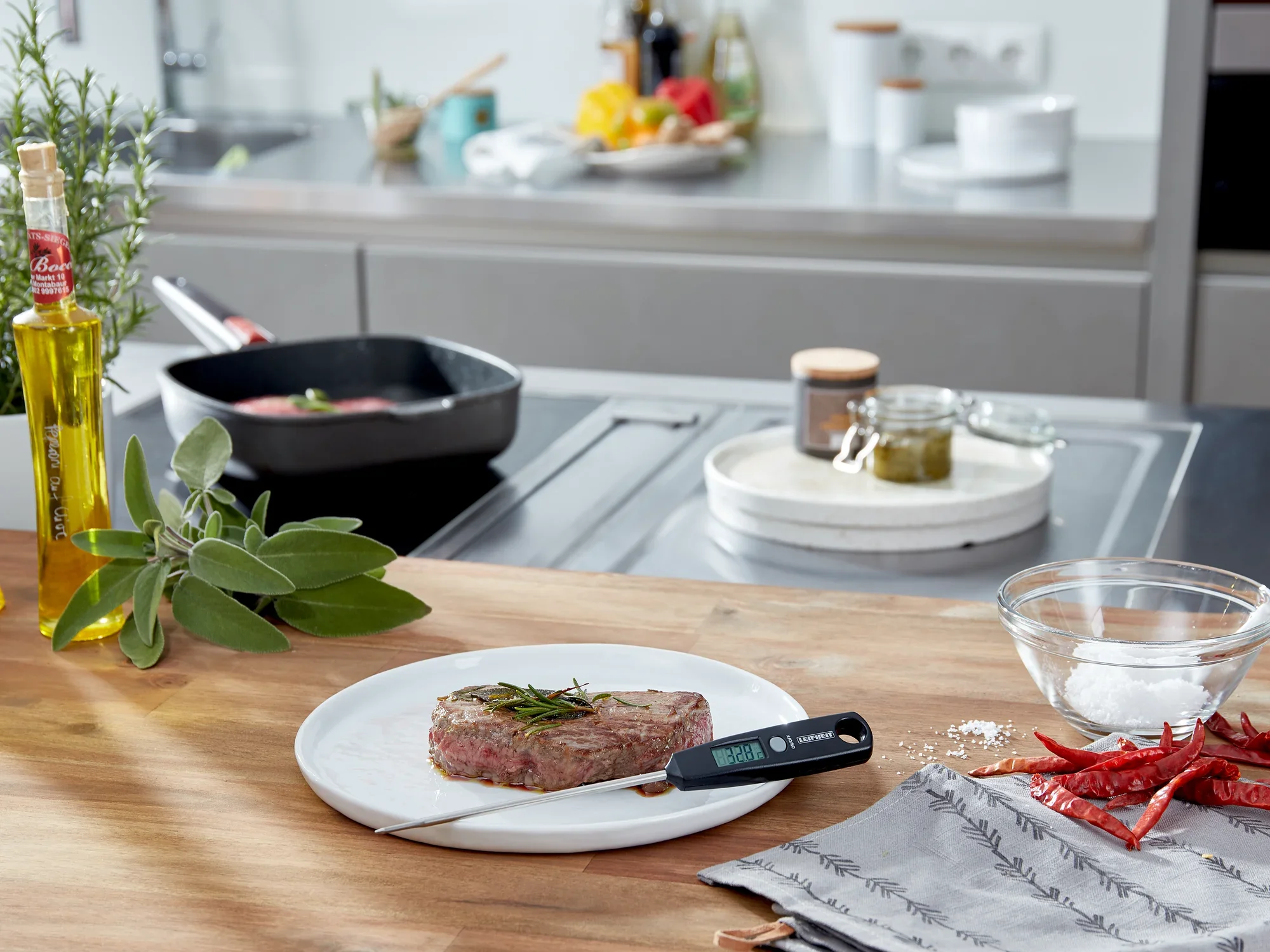 Uniwersalny termometr cyfrowy leży na talerzu ze stekiem w nowoczesnej kuchni. W tle widoczne są przybory kuchenne i przyprawy.