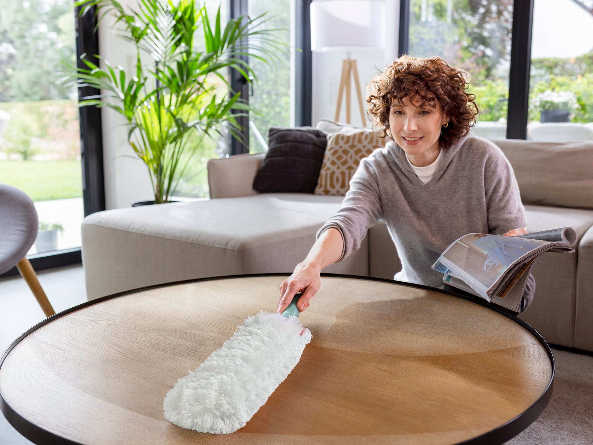 Eine Person benutzt den SUPERDUSTER XL Staubwedel, um einen runden Holztisch in einem modernen Wohnzimmer zu reinigen. Der Staubwedel hat einen weißen, flauschigen Kopf, ideal für sanfte Reinigung.