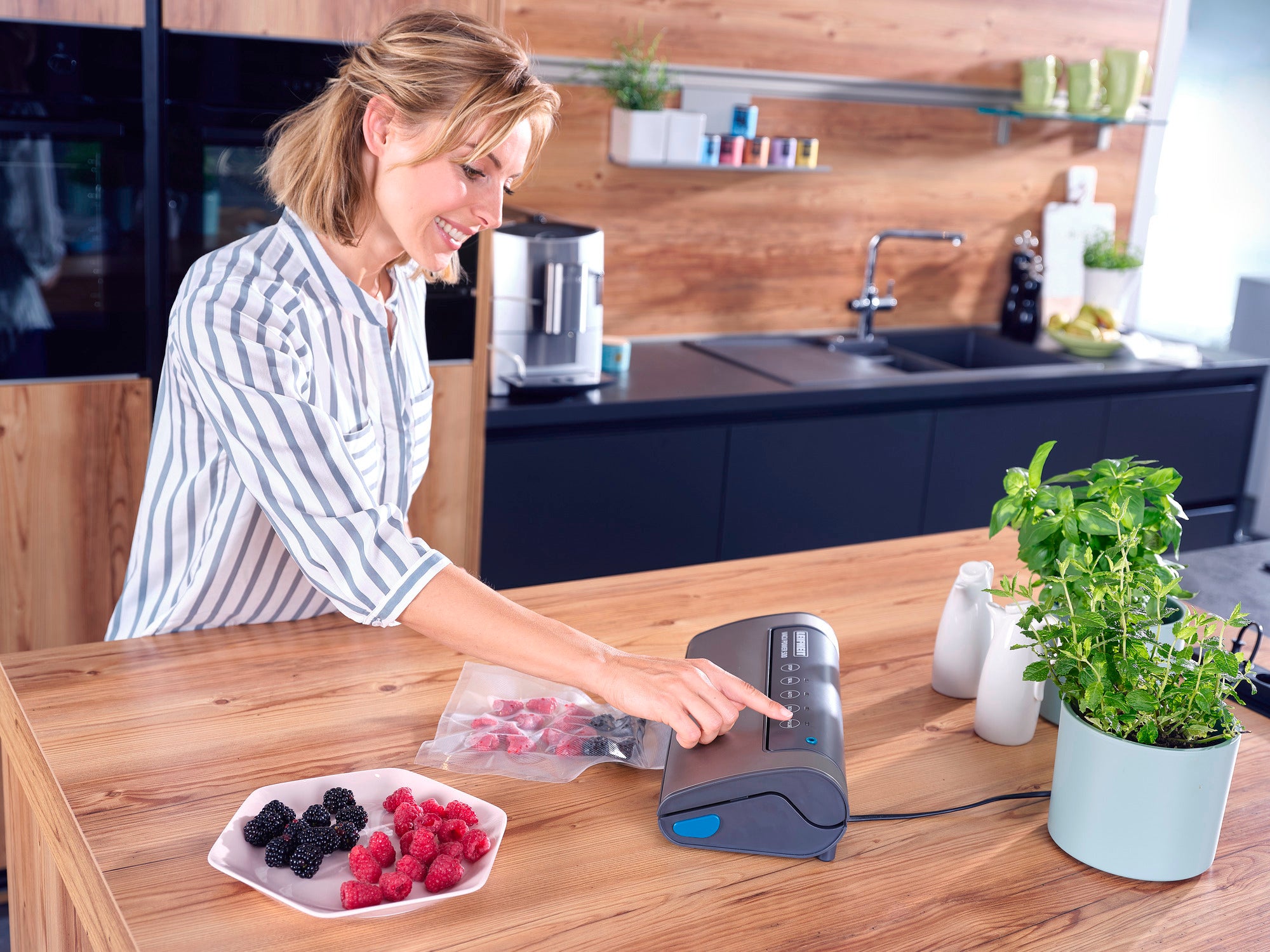 Eine Frau benutzt das Vakuumiergerät Vacu Power 500 in einer modernen Küche. Sie vakuumiert Beeren in einem Beutel. Auf dem Tisch stehen frische Kräuter und eine Schale mit Beeren.