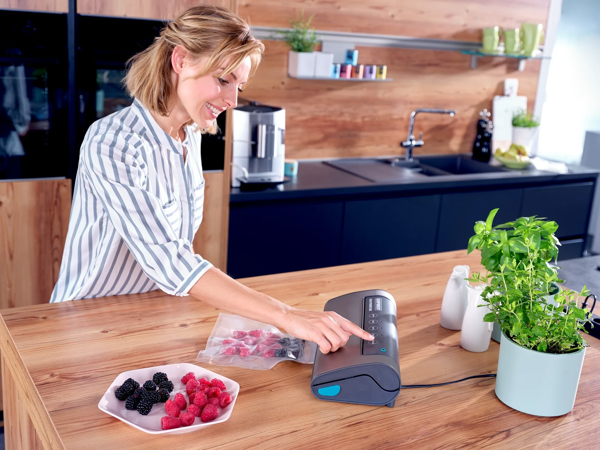Kobieta używa zgrzewarki próżniowej Vacu Power 500 w nowoczesnej kuchni. Uszczelnia próżniowo jagody w woreczku. Na stole leżą świeże zioła i miska z jagodami.