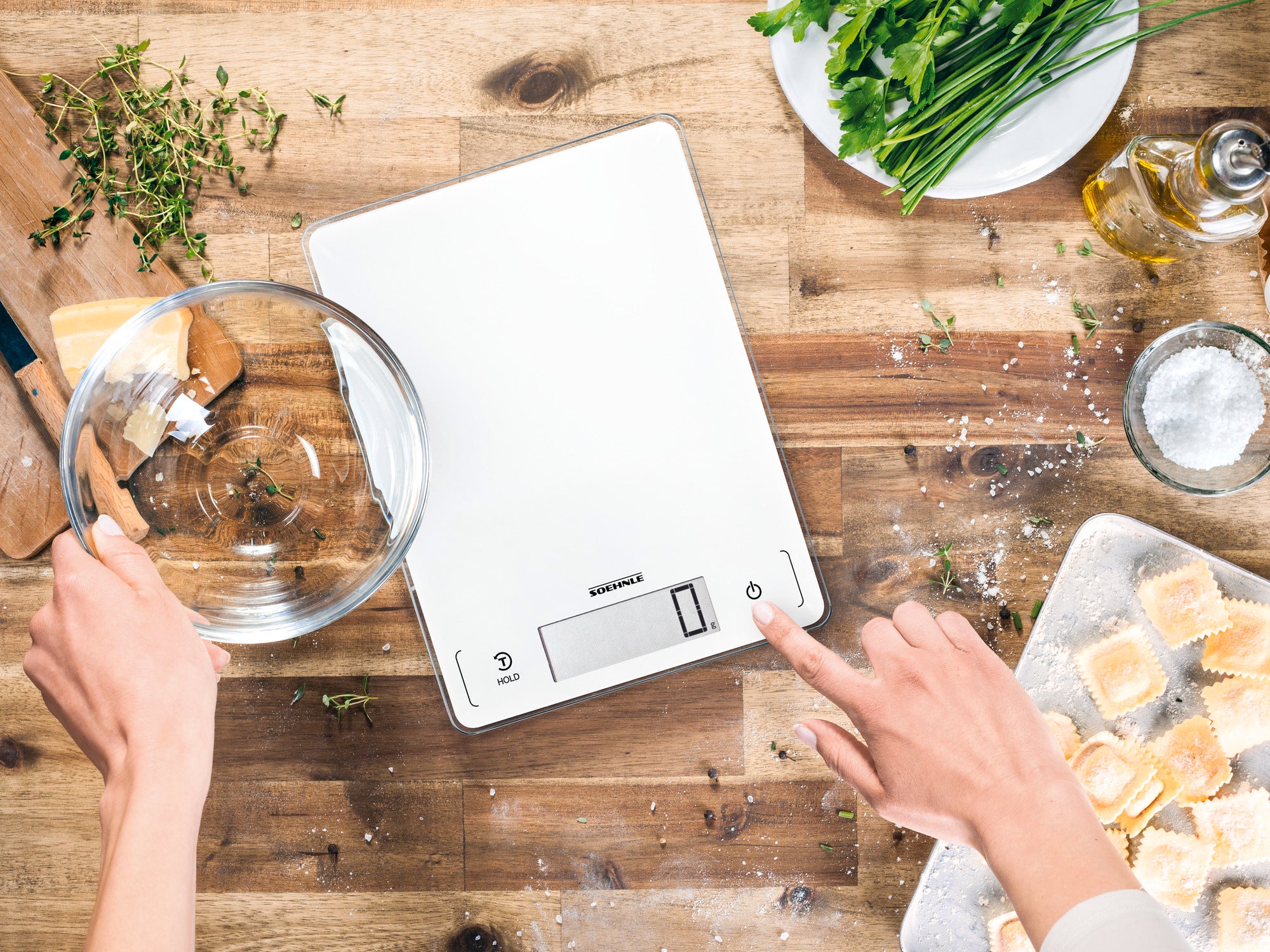 Balance de cuisine numérique Page Profi 300 sur une table en bois, entourée d'herbes fraîches, d'huile et de pâtes. Une main tient un saladier tandis que l'autre actionne la balance.