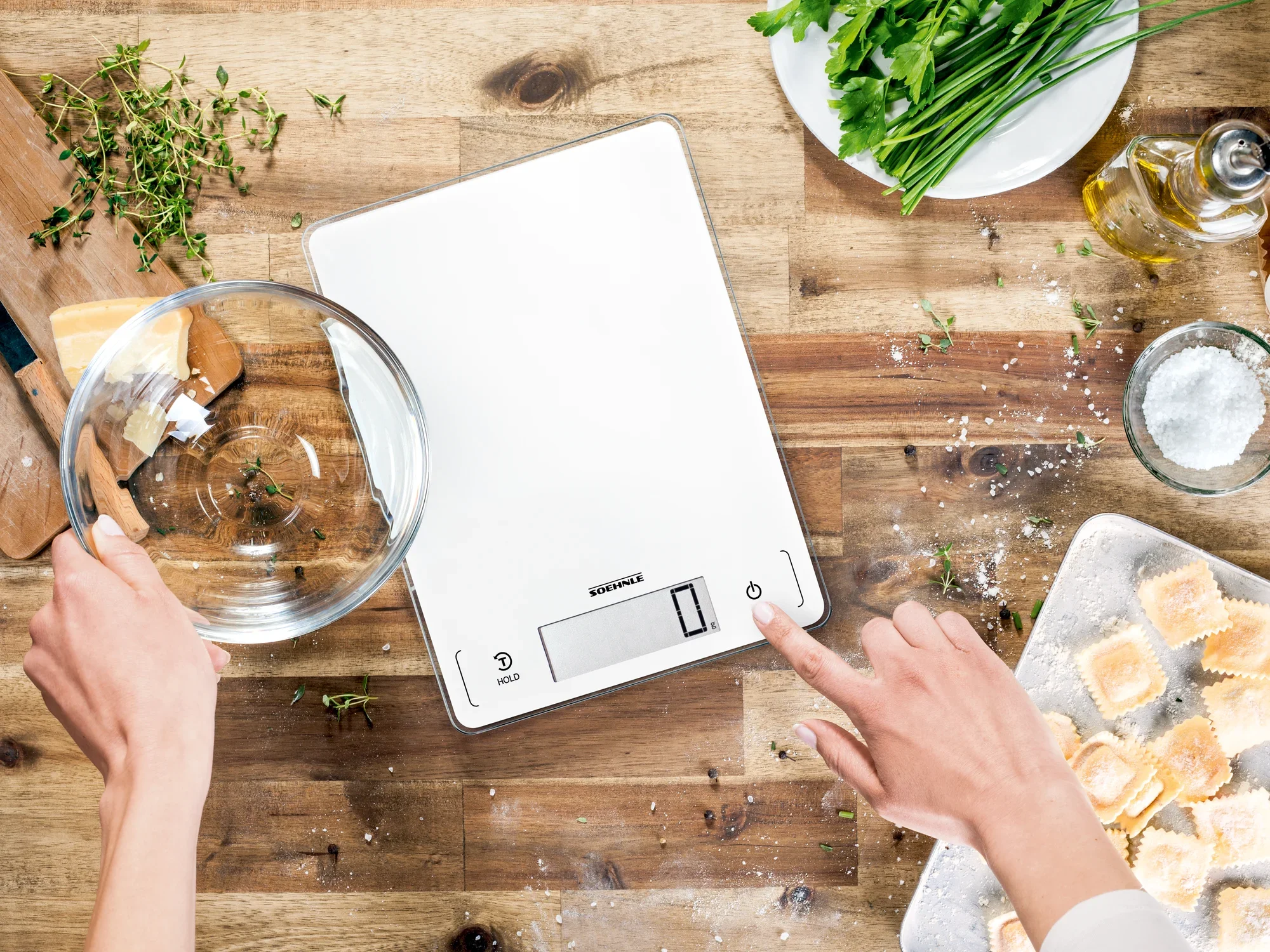 Digitale Küchenwaage Page Profi 300 auf einem Holztisch, umgeben von frischen Kräutern, Öl und Teigwaren. Eine Hand hält eine Schüssel, während die andere die Waage bedient.